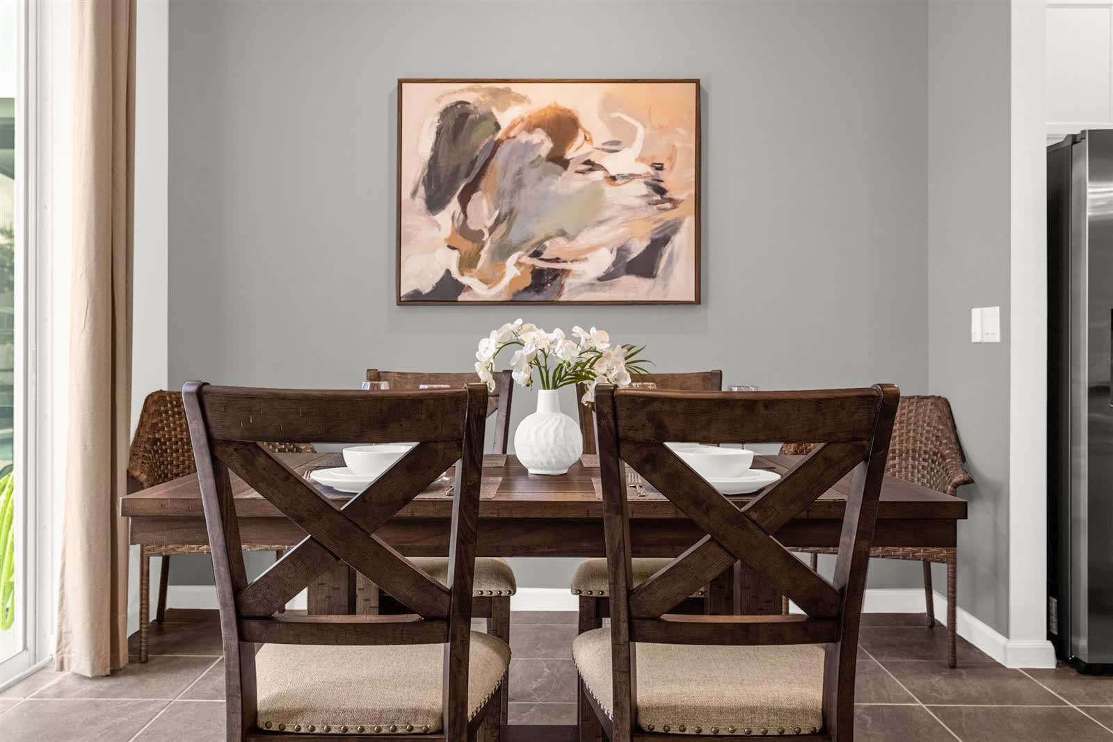 A dining room features a dark wood rectangular table with six matching chairs, set against a light gray wall with a large abstract painting. White dinnerware and wine glasses are visible on the table, with a white vase of orchids as a centerpiece.