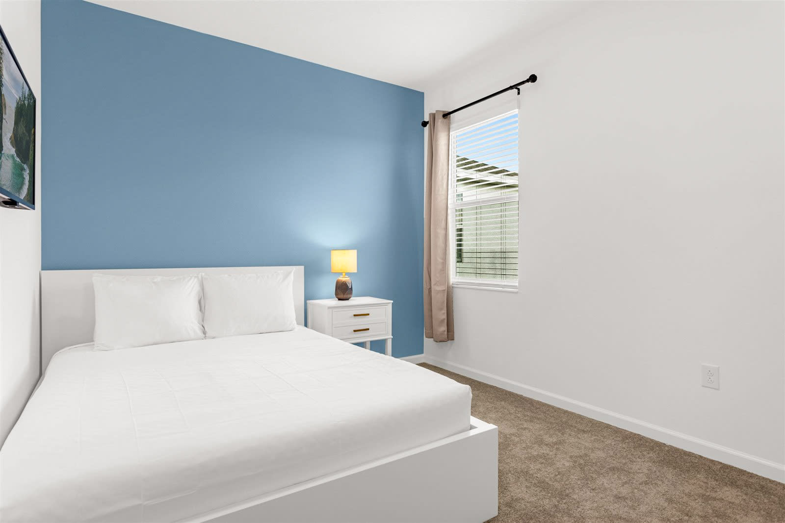 A minimalist bedroom with a white bed and white linens, a white nightstand with a yellow lamp, and a blue accent wall. A window with blinds and a curtain is visible on the right, offering a glimpse of the exterior.