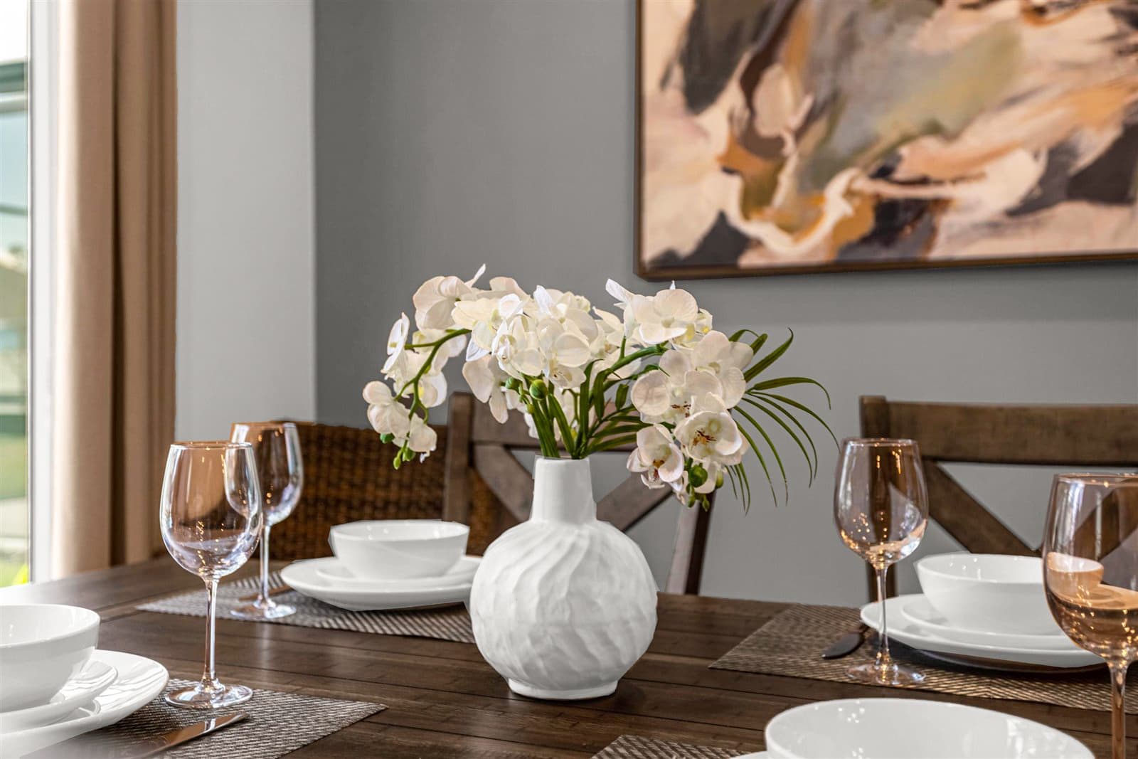 Close-up view of a dark wood dining table set with white dinnerware, wine glasses, and a white vase holding white orchids. The table is surrounded by dark wood chairs with upholstered seats, and an abstract painting hangs on the gray wall in the background.