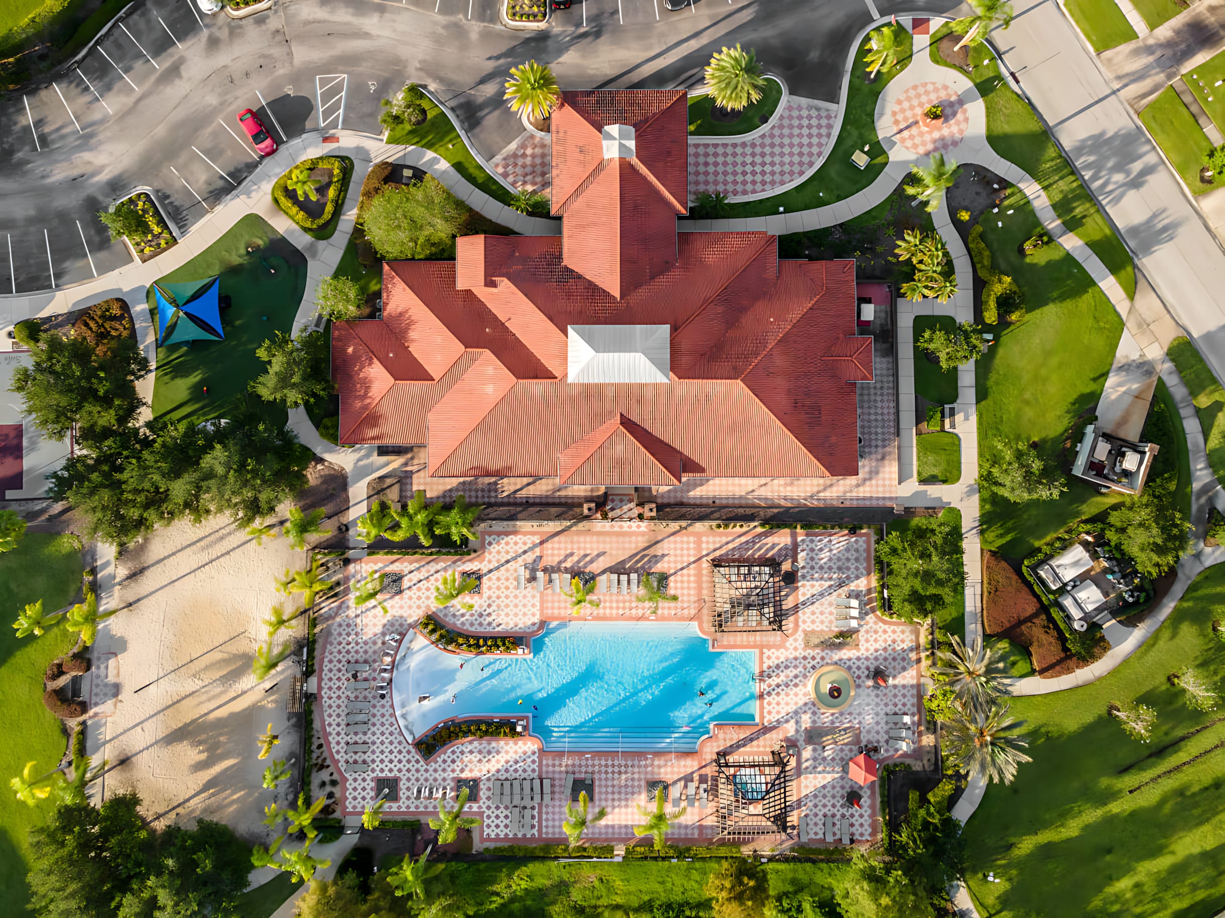 An aerial view of a large resort complex featuring a central building with a red tile roof, a swimming pool with lounge areas, and surrounding residential buildings. The property is set amidst green landscaping and parking lots.