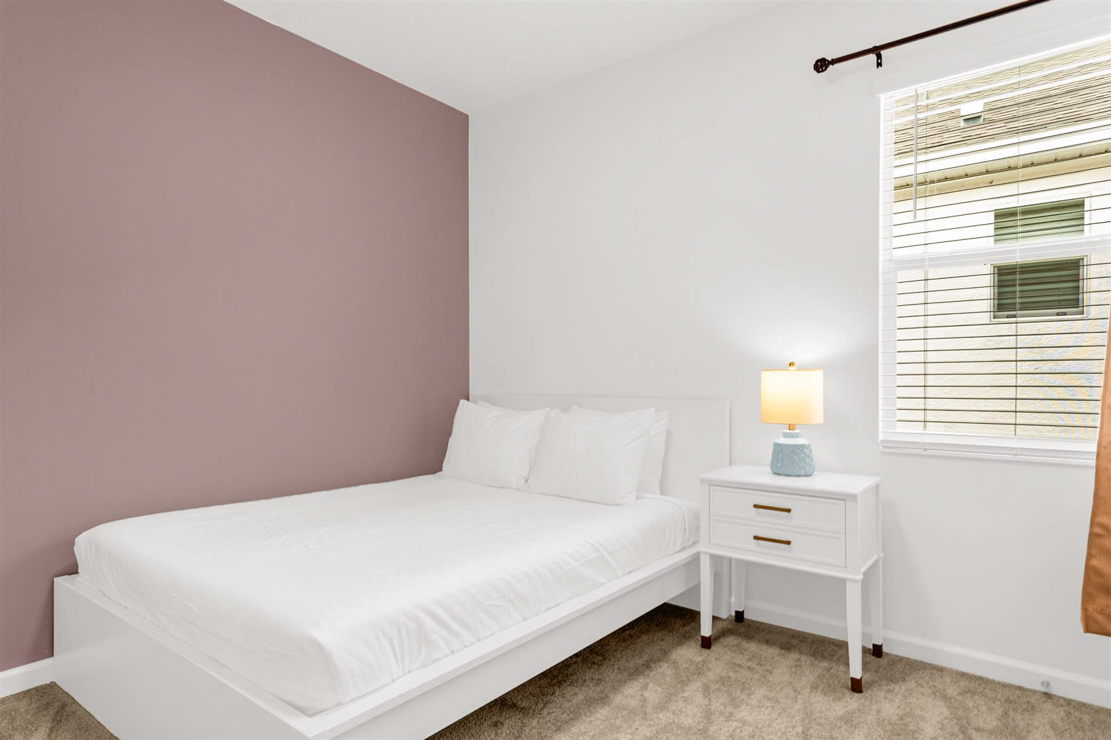 A minimalist bedroom with a white bed and white linens, a white nightstand with a blue and white lamp, and a muted purple accent wall. A window with blinds and a curtain is visible on the right.