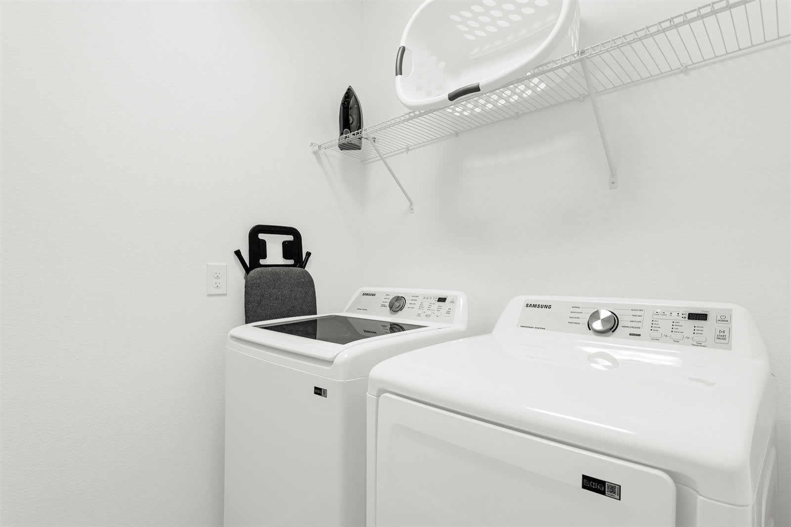 A functional laundry room equipped with a white top-load washing machine and a white front-load dryer. A wire shelf above holds a laundry basket and an iron, with an ironing board leaning against the wall.