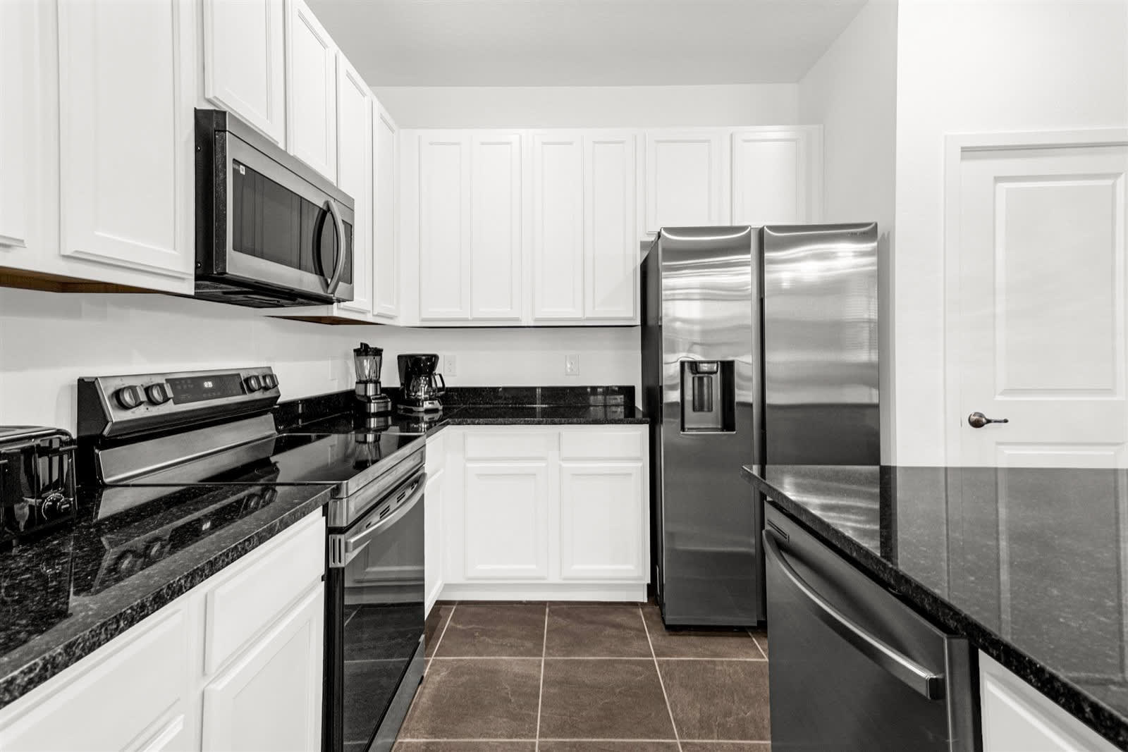 A close-up view of a modern kitchen showing white shaker cabinets, black granite countertops, and stainless steel appliances like a range, microwave, and dishwasher. A coffee maker and toaster are visible on the counter, with dark tile flooring.