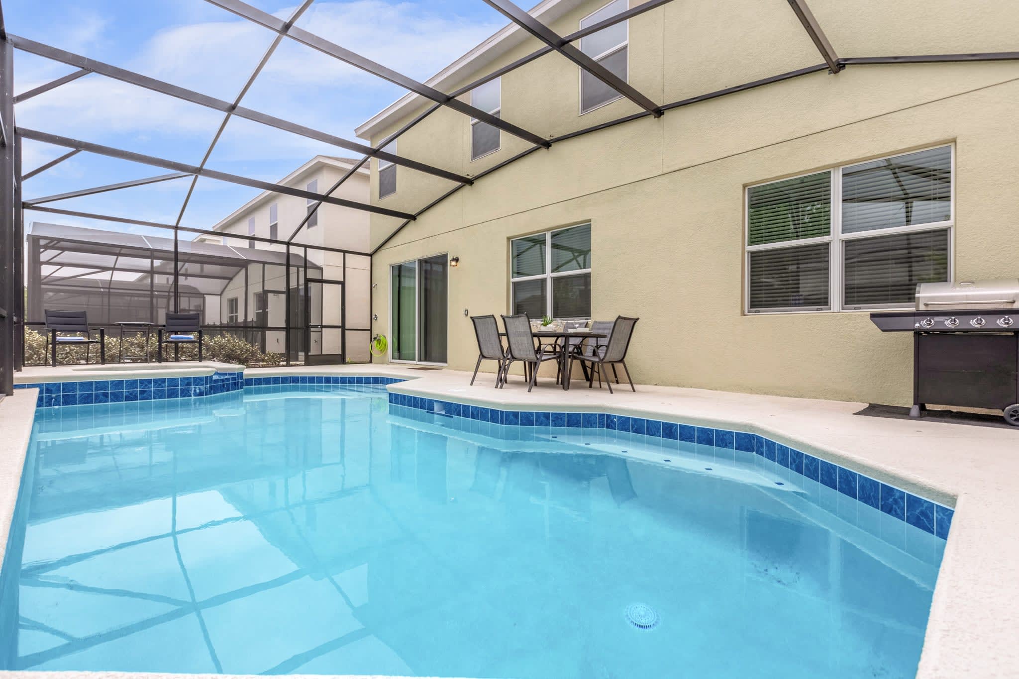 A wide view of the screened outdoor pool area shows a rectangular swimming pool with blue tile accents and a light concrete deck. The beige house exterior with multiple windows and a sliding glass door is visible, along with an outdoor dining set and a grill.