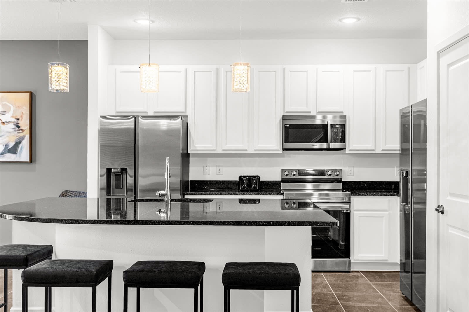 A modern kitchen features white shaker cabinets, black granite countertops, and stainless steel appliances, including a double-door refrigerator and a range. A large kitchen island with a sink and three bar stools is in the foreground, with decorative pendant lights above.
