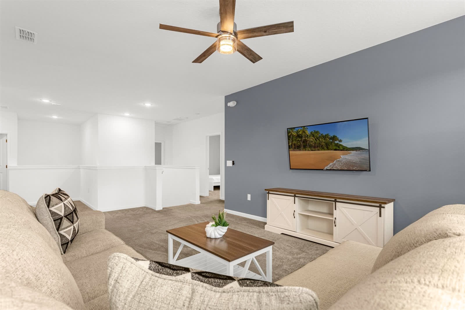 An upstairs living area features a large beige sectional sofa with patterned throw pillows, a wooden coffee table, and a wall-mounted TV on a gray accent wall. A ceiling fan with wooden blades and a light fixture is installed overhead, with carpeted flooring.