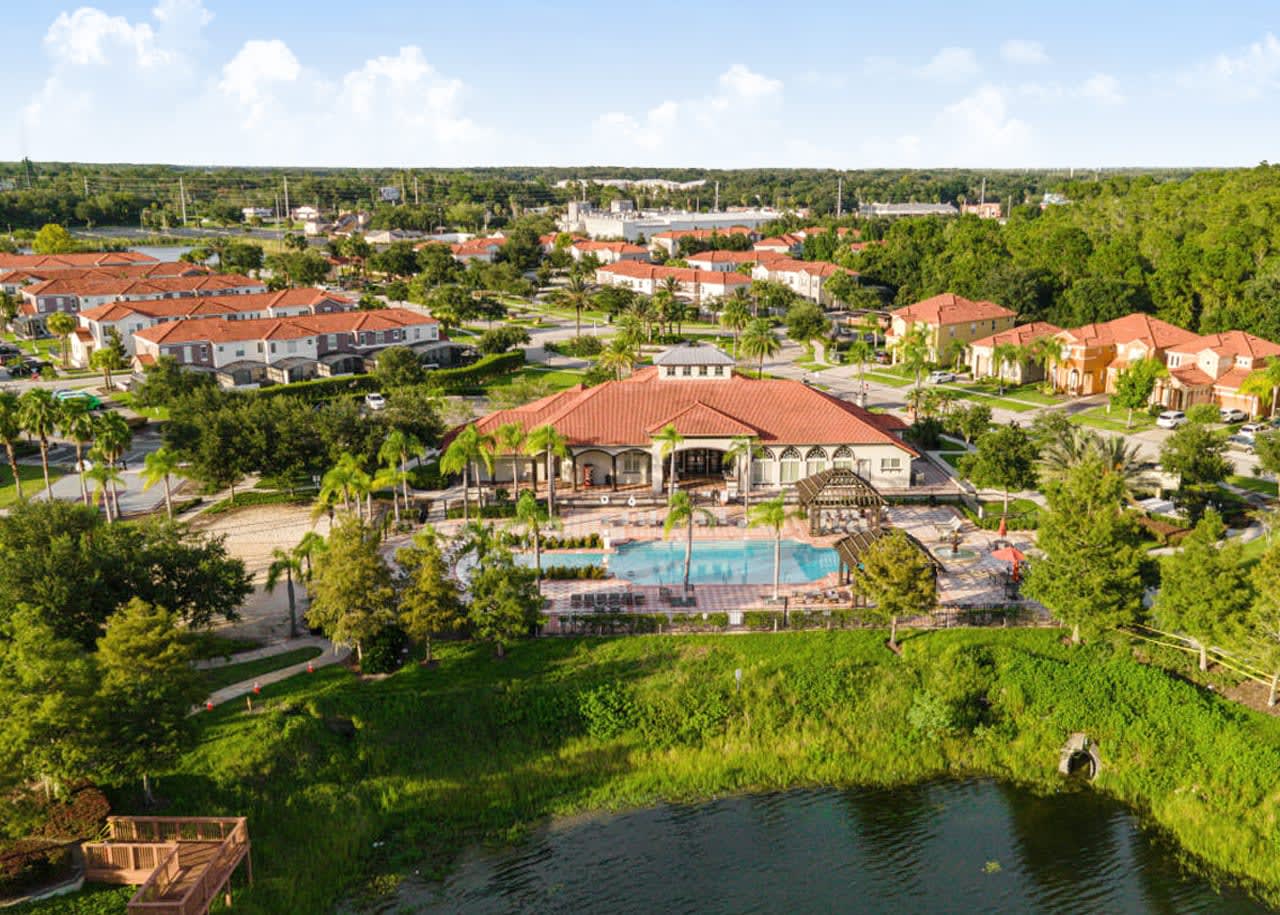 A wide aerial shot of a resort community, showcasing a large central building with a red tile roof and an adjacent swimming pool area. The community is surrounded by numerous residential buildings, green spaces, and a body of water in the foreground.