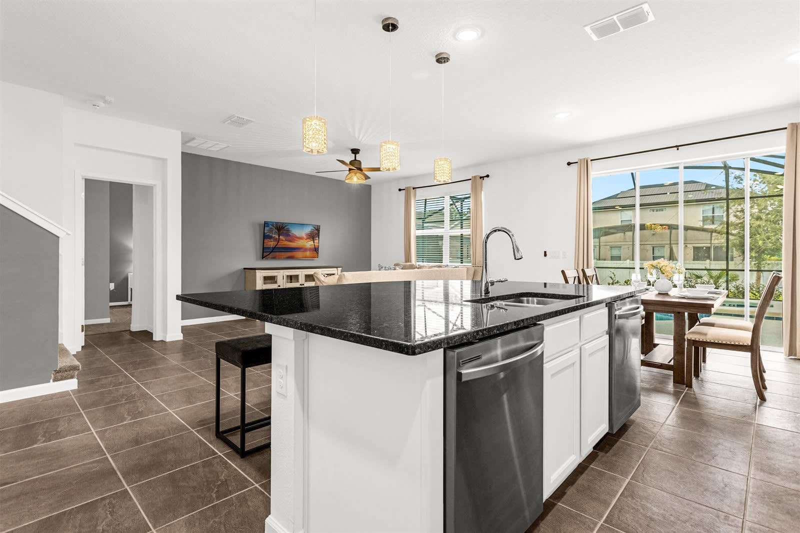 Open concept view of a modern kitchen with white cabinetry, black granite countertops, and stainless steel appliances, including a large island with a sink and bar seating. The dining area is visible through a large sliding glass door leading to an outdoor pool area.