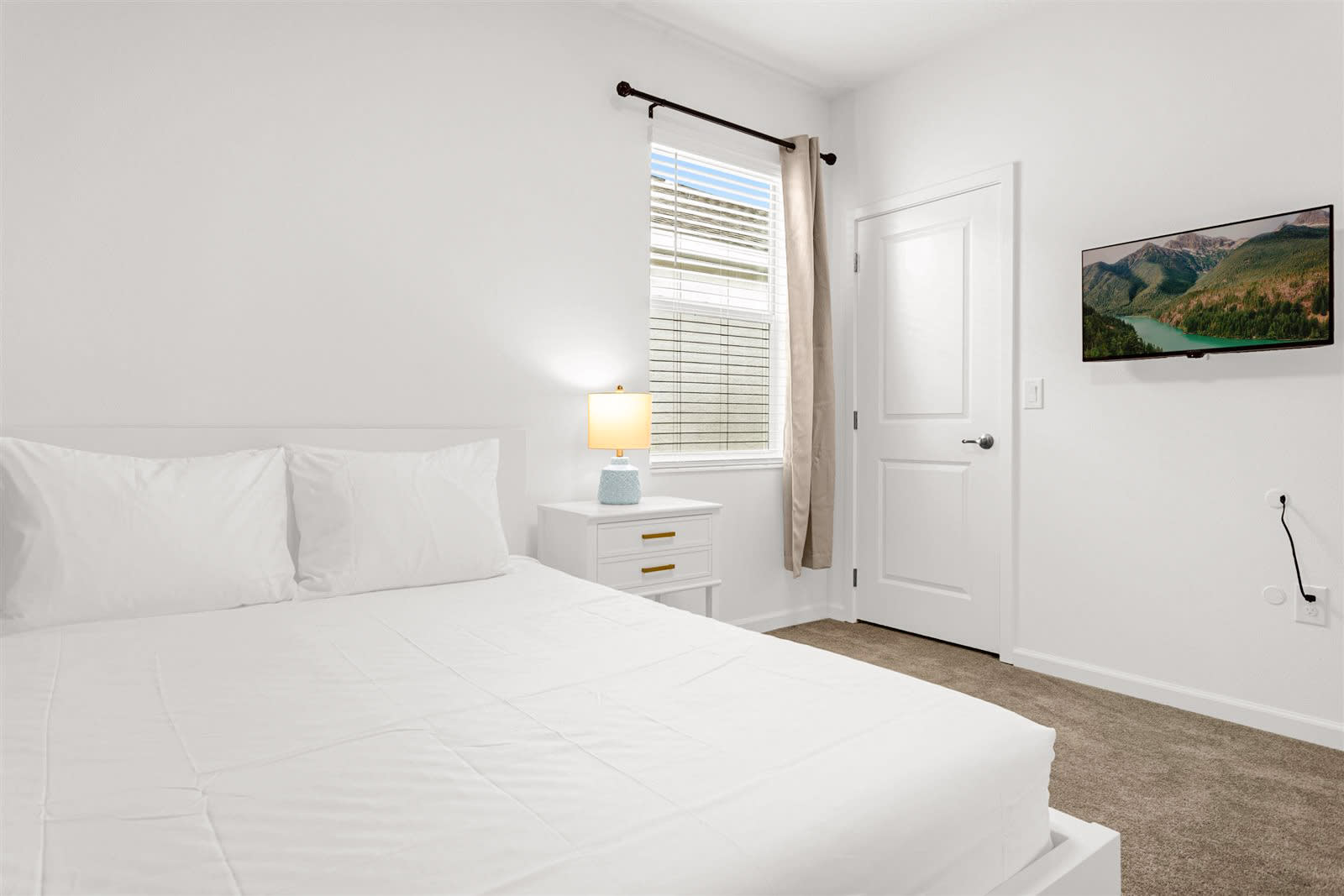 A bright bedroom with a white bed and white linens, a white nightstand with a blue and white lamp, and a wall-mounted TV. A window with blinds and a curtain is on the left, and a white door is visible on the right.
