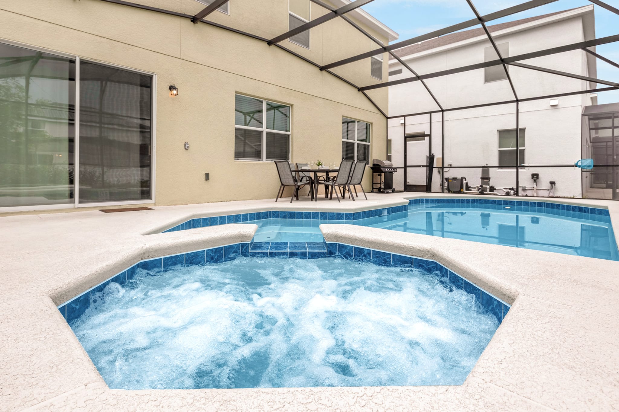 An outdoor screened pool area features a rectangular swimming pool and an adjacent bubbling hot tub, both with blue tile accents. A small outdoor dining set with four chairs and a grill are visible on the light concrete deck next to the beige-colored house.