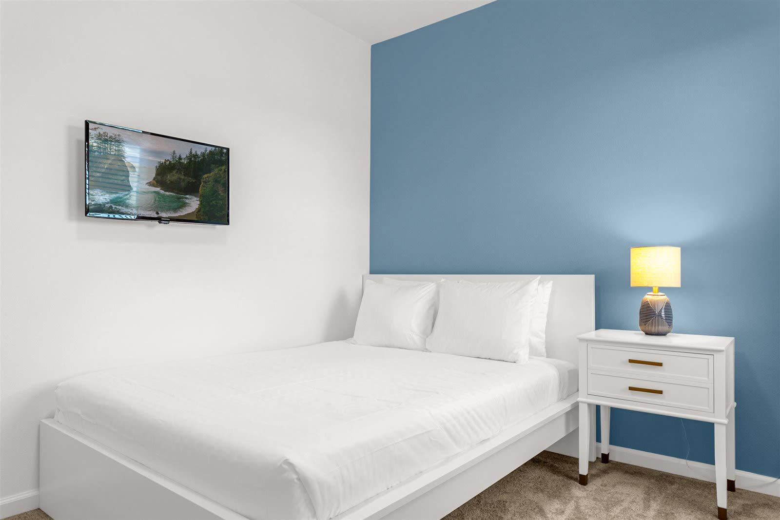 A minimalist bedroom featuring a white bed with white linens, a wall-mounted TV displaying a scenic landscape, and a striking blue accent wall contrasting with white walls. A white nightstand with a yellow lamp is next to the bed.
