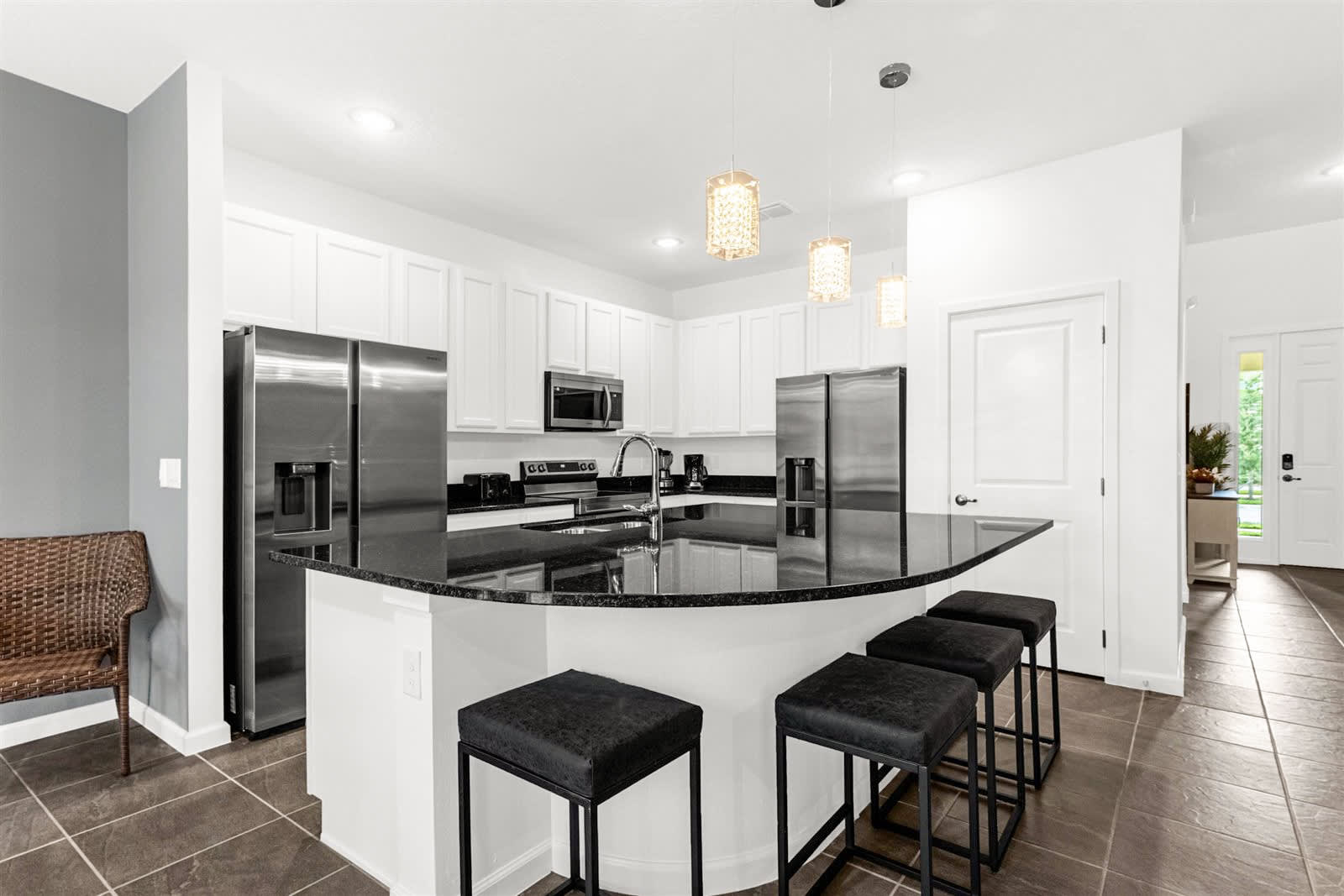 A bright kitchen with white shaker cabinets, black granite countertops, and stainless steel appliances, including a large double-door refrigerator. A curved kitchen island with bar seating and decorative pendant lights overhead defines the space.