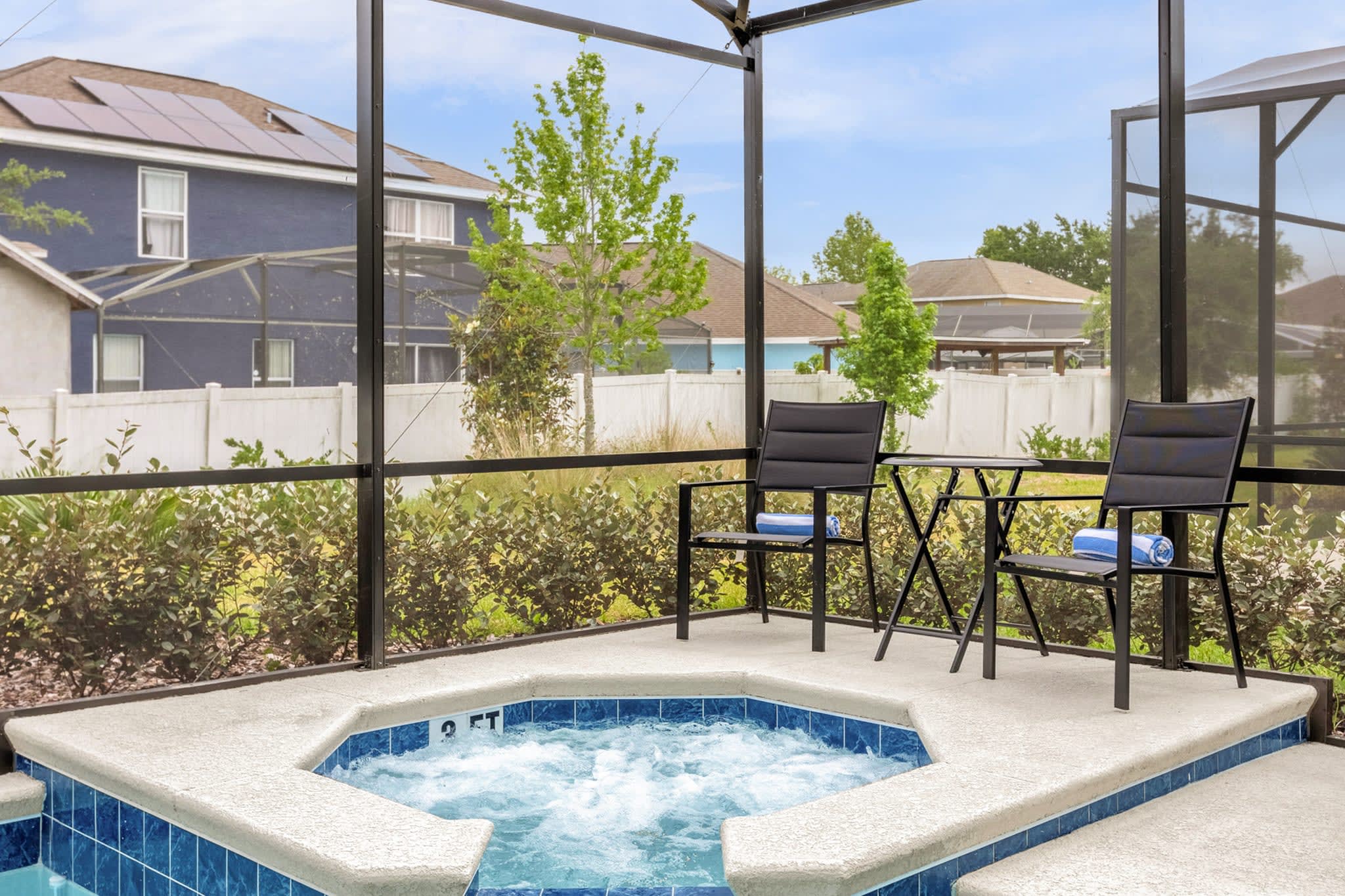 A close-up view of the bubbling hot tub, integrated into the pool deck and enclosed by a black screen. Two black outdoor chairs and a small table are placed on the deck next to the hot tub, with lush green landscaping and a white fence in the background.
