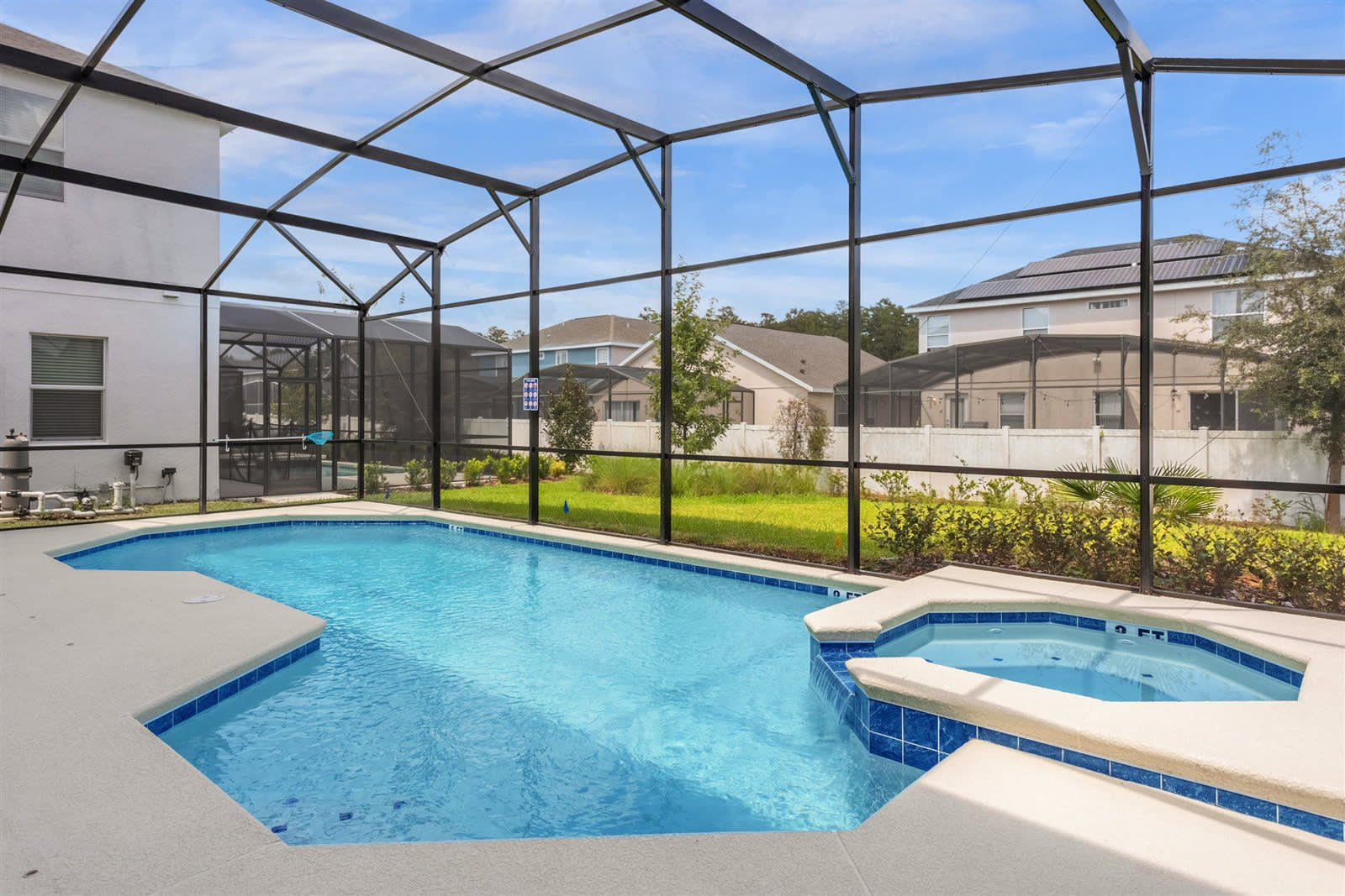 An outdoor swimming pool and integrated hot tub are enclosed within a black screened cage, surrounded by a light-colored concrete deck and a small strip of green grass and shrubs. In the background, other houses with screened pools are visible under a blue sky.