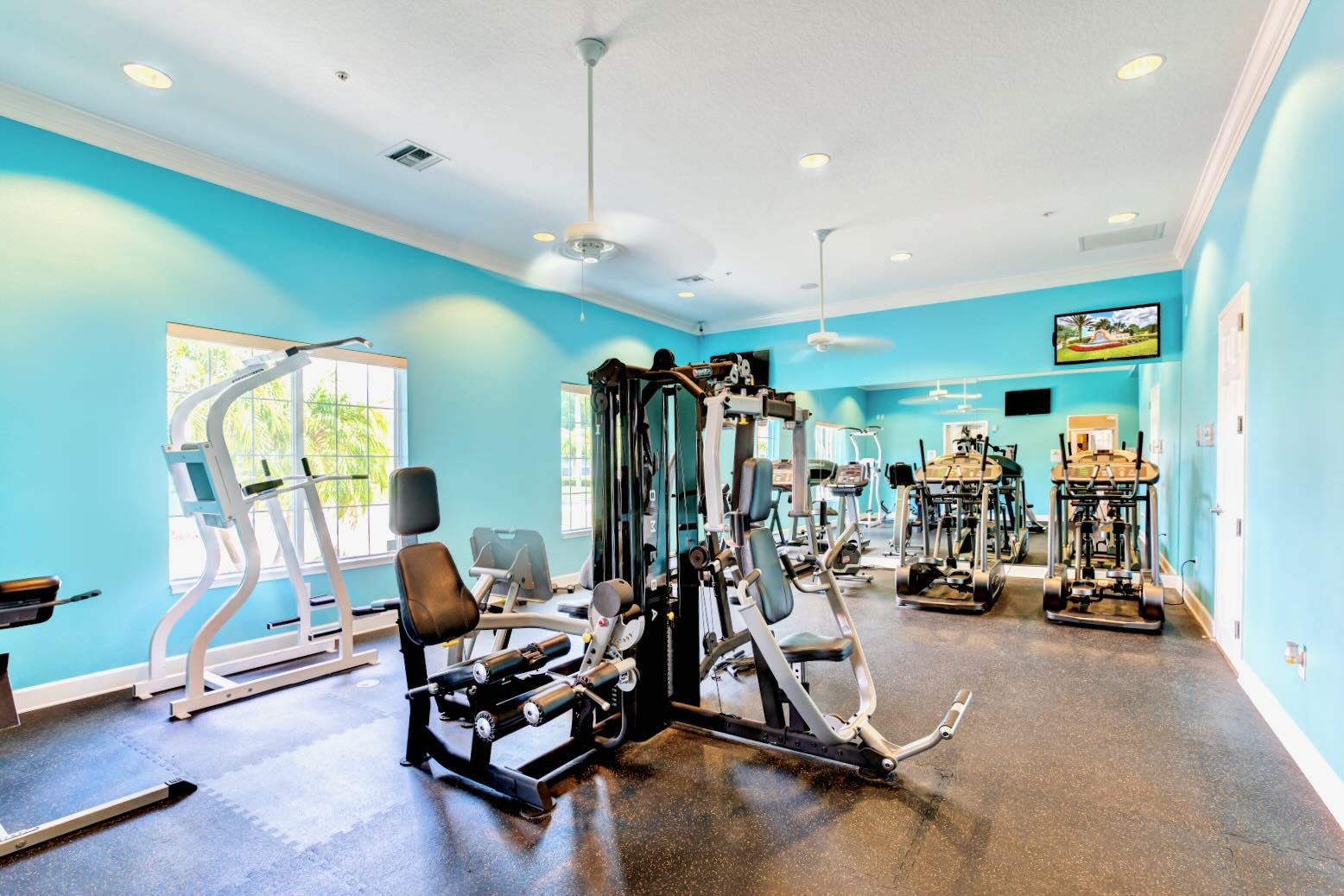 Bright and spacious fitness center with light blue walls, featuring a variety of exercise equipment including treadmills, ellipticals, and weight machines, with large windows and ceiling fans.