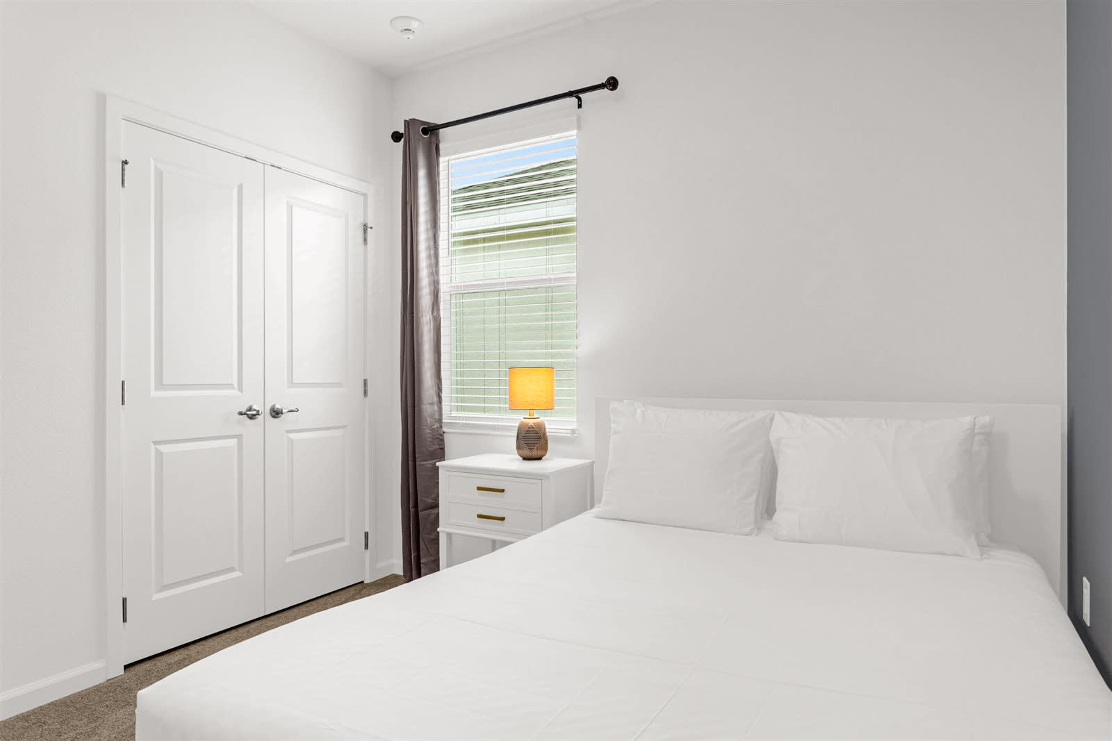 View of a minimalist bedroom showing a white platform bed, a single white nightstand with gold pulls, and a window with blinds and drapes, with a grey accent wall and white closet doors.