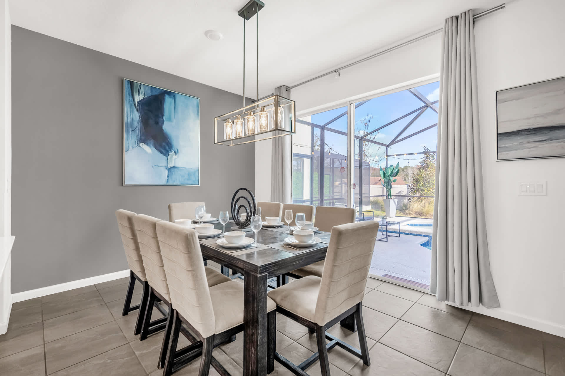 A stylish dining area features a large dark wood dining table with eight upholstered chairs, illuminated by a modern rectangular chandelier. A dark gray accent wall with abstract art complements the space, which offers a view of the outdoor pool through sliding glass doors.