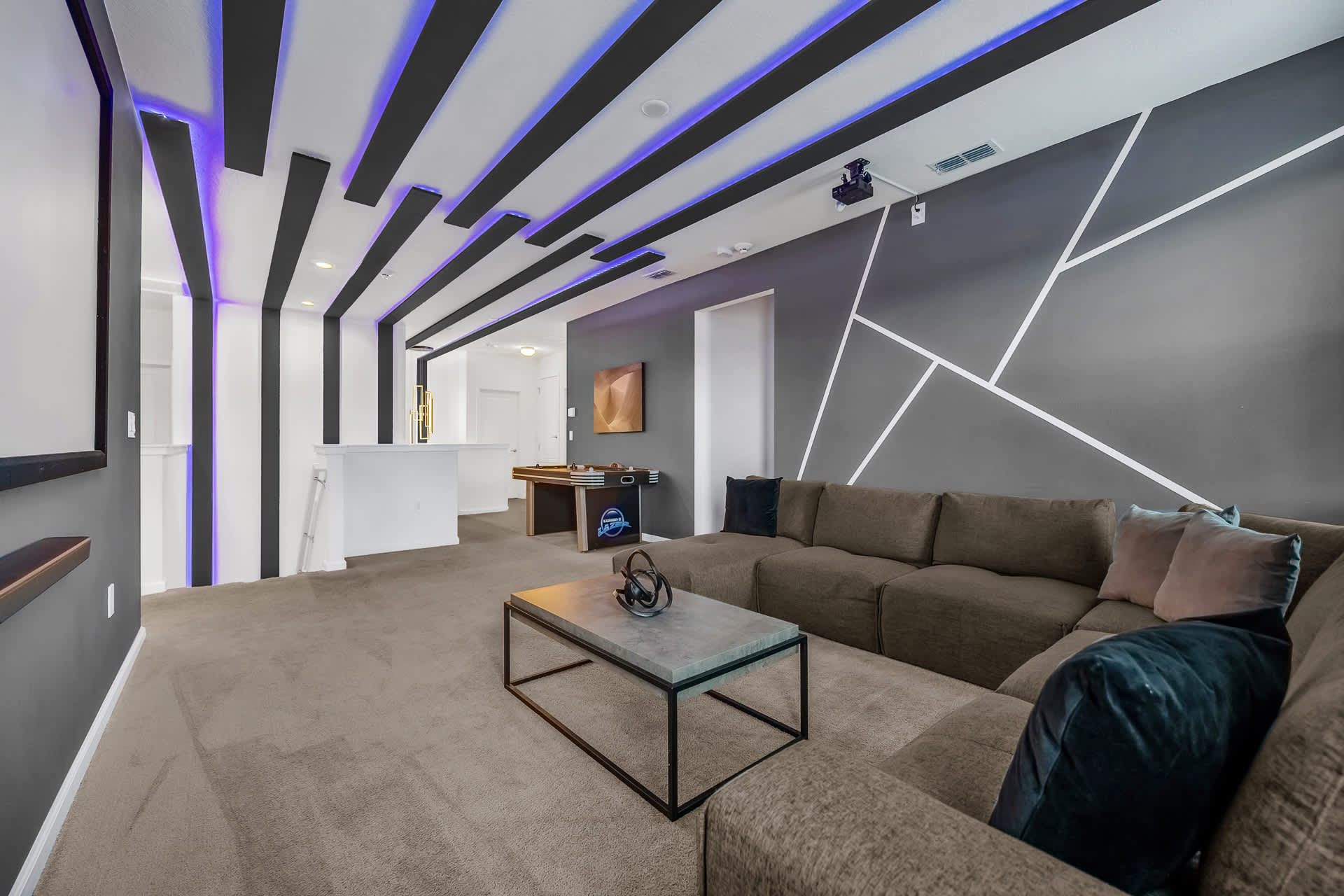 A spacious game and media room features a large L-shaped sectional sofa, a modern coffee table, and an air hockey table in the background. The room boasts a dark gray accent wall with geometric white lines and a ceiling with black beams and purple LED lighting.