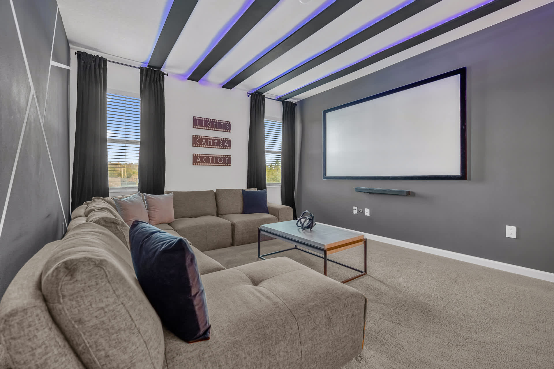 A modern media room features a large projector screen on a dark gray wall, a comfortable L-shaped sectional sofa, and a coffee table. The ceiling is adorned with black beams illuminated by purple LED lights, creating a cinematic ambiance.