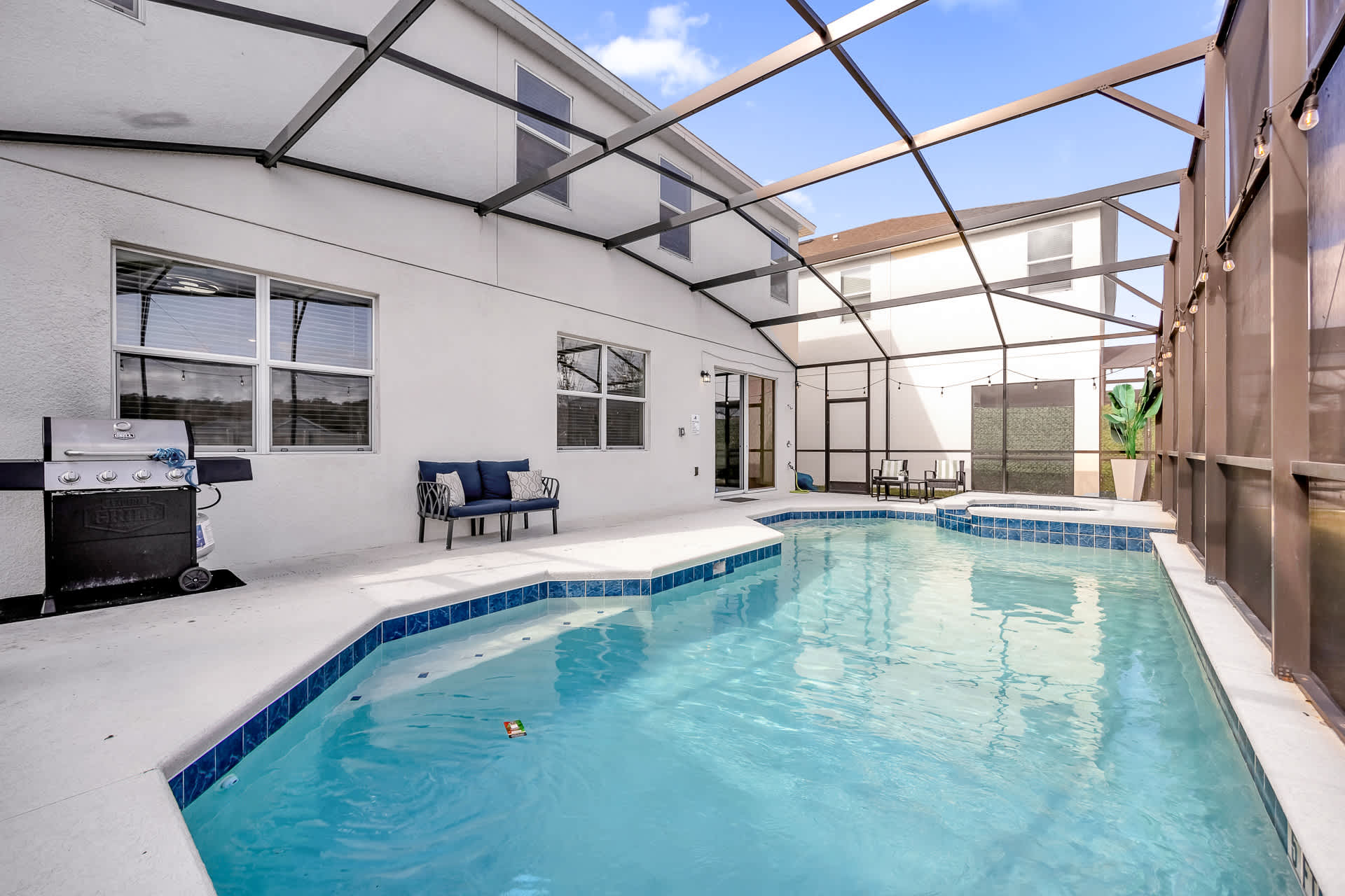 A bright, daytime view of the screened-in pool area, featuring a large swimming pool with an integrated hot tub, concrete decking, and string lights, with a BBQ grill and outdoor bench visible.