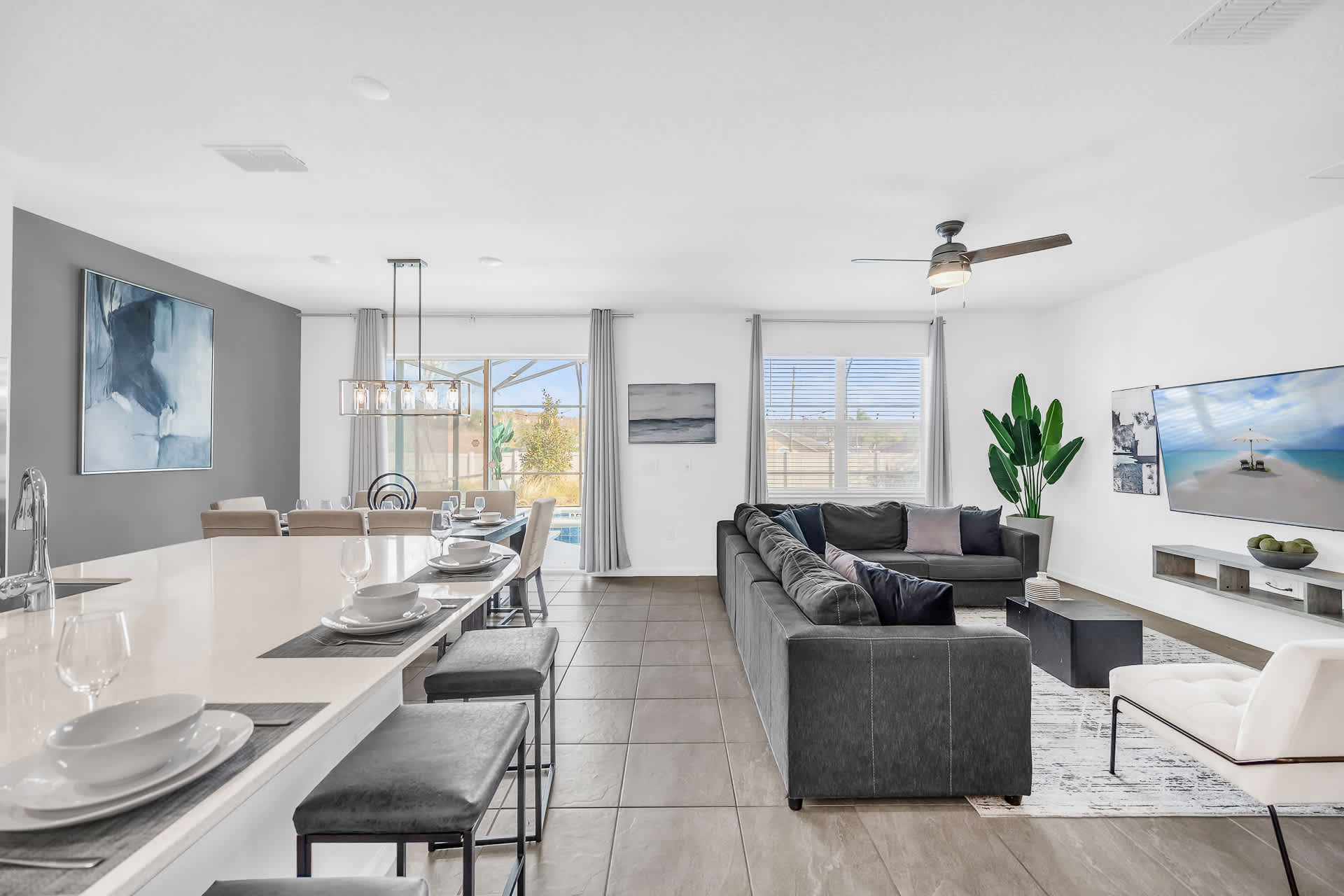 A spacious open-concept living area encompasses a comfortable living room with a sectional sofa, a large kitchen island with seating, and a dining area with a view of the outdoor pool. The room is brightly lit with natural light from large windows and sliding glass doors.