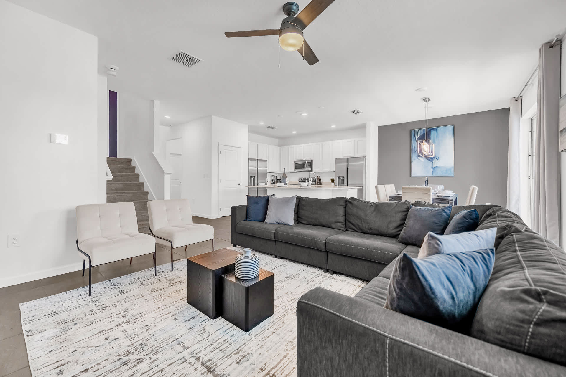 An open-concept living area features a large dark gray sectional sofa, two accent chairs, and a modern coffee table on a patterned area rug. The space flows into the kitchen and dining areas, with stairs leading to the upper floor.