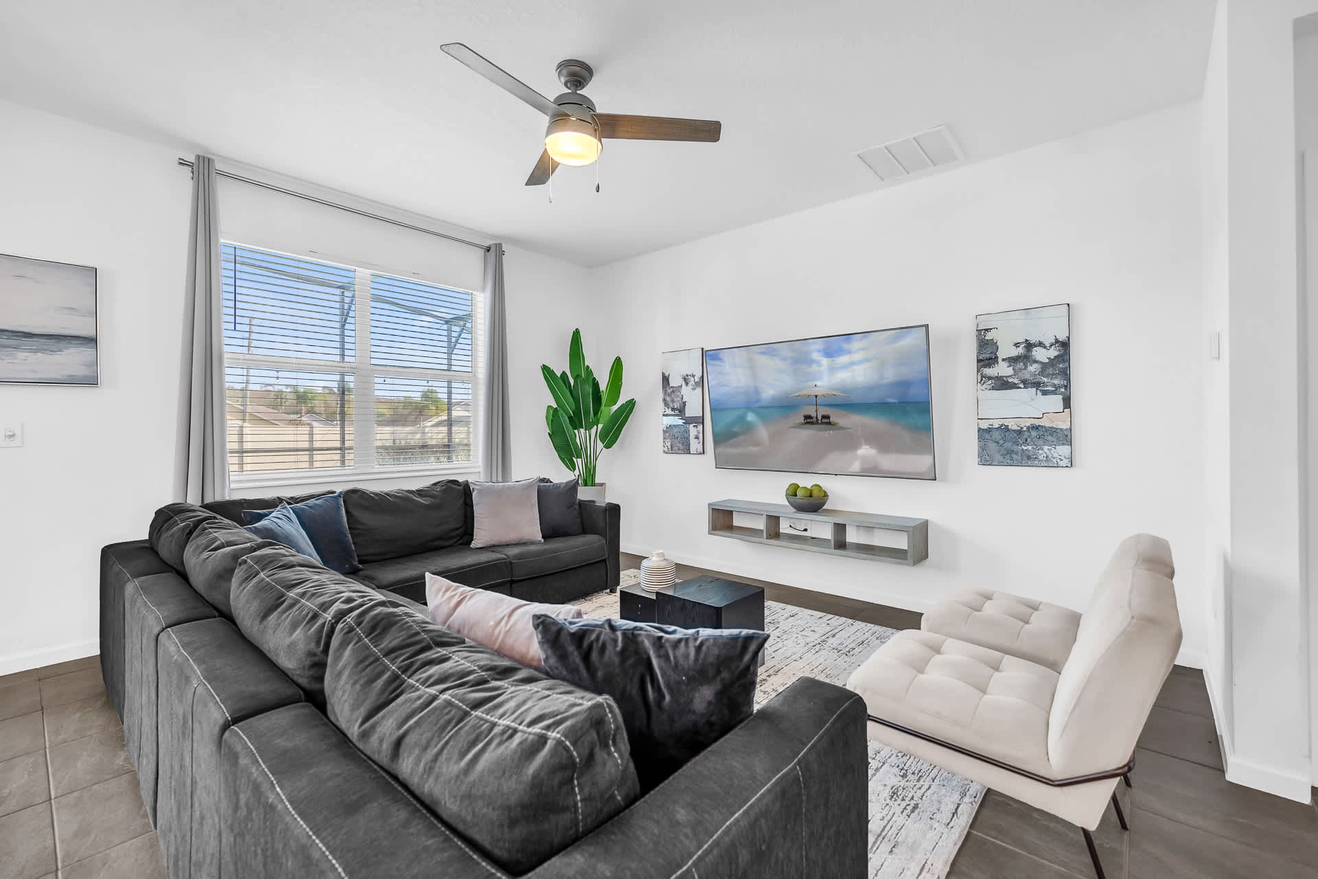 A bright living room features a large dark gray sectional sofa with colorful throw pillows, facing a wall-mounted flat-screen TV flanked by abstract art. A modern ceiling fan and a large window with blinds complete the space.