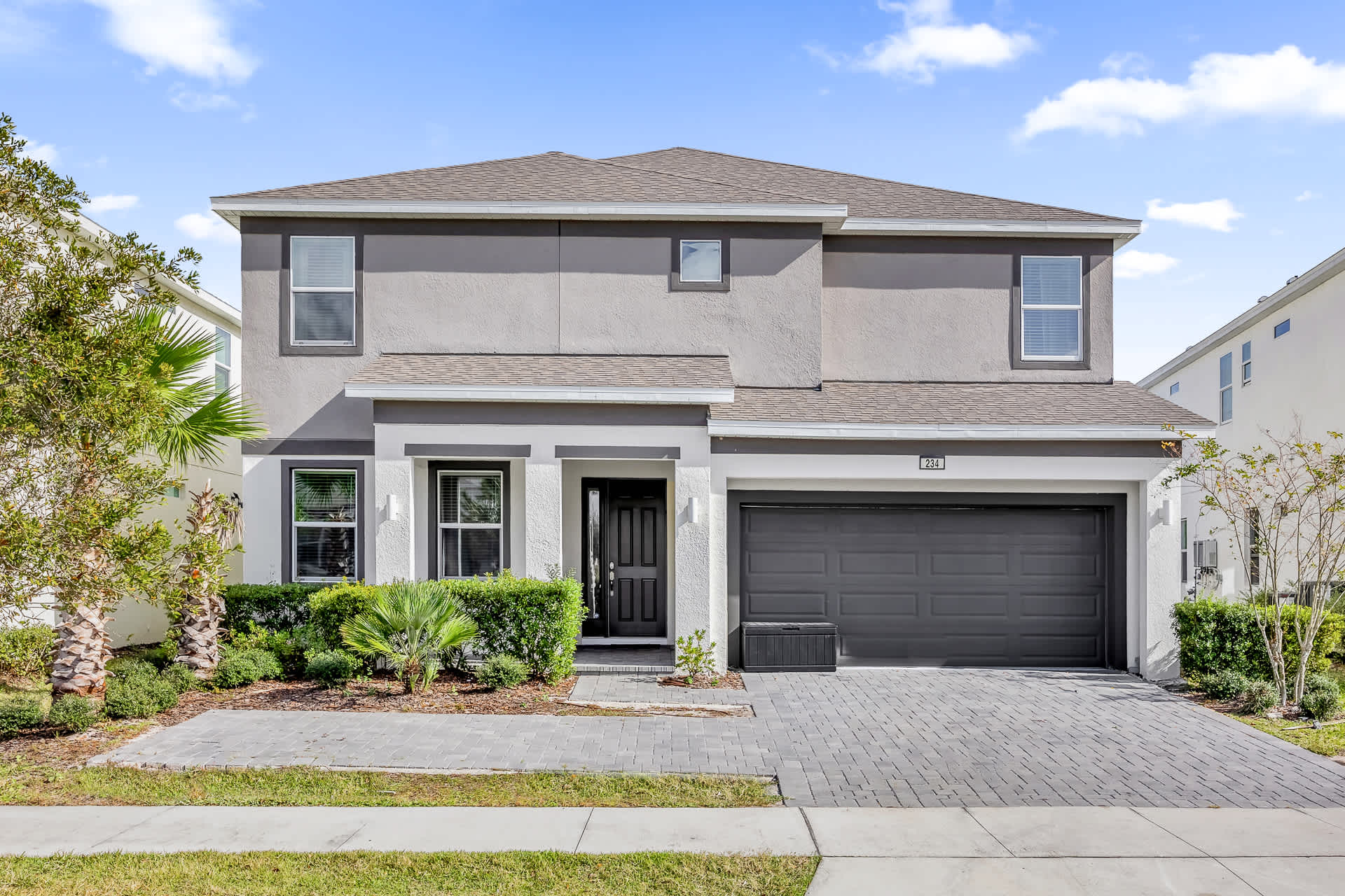 Modern two-story house with a gray and white stucco exterior, a dark gray garage door, and a paved driveway. Lush green landscaping with bushes and small palm trees frames the entrance.
