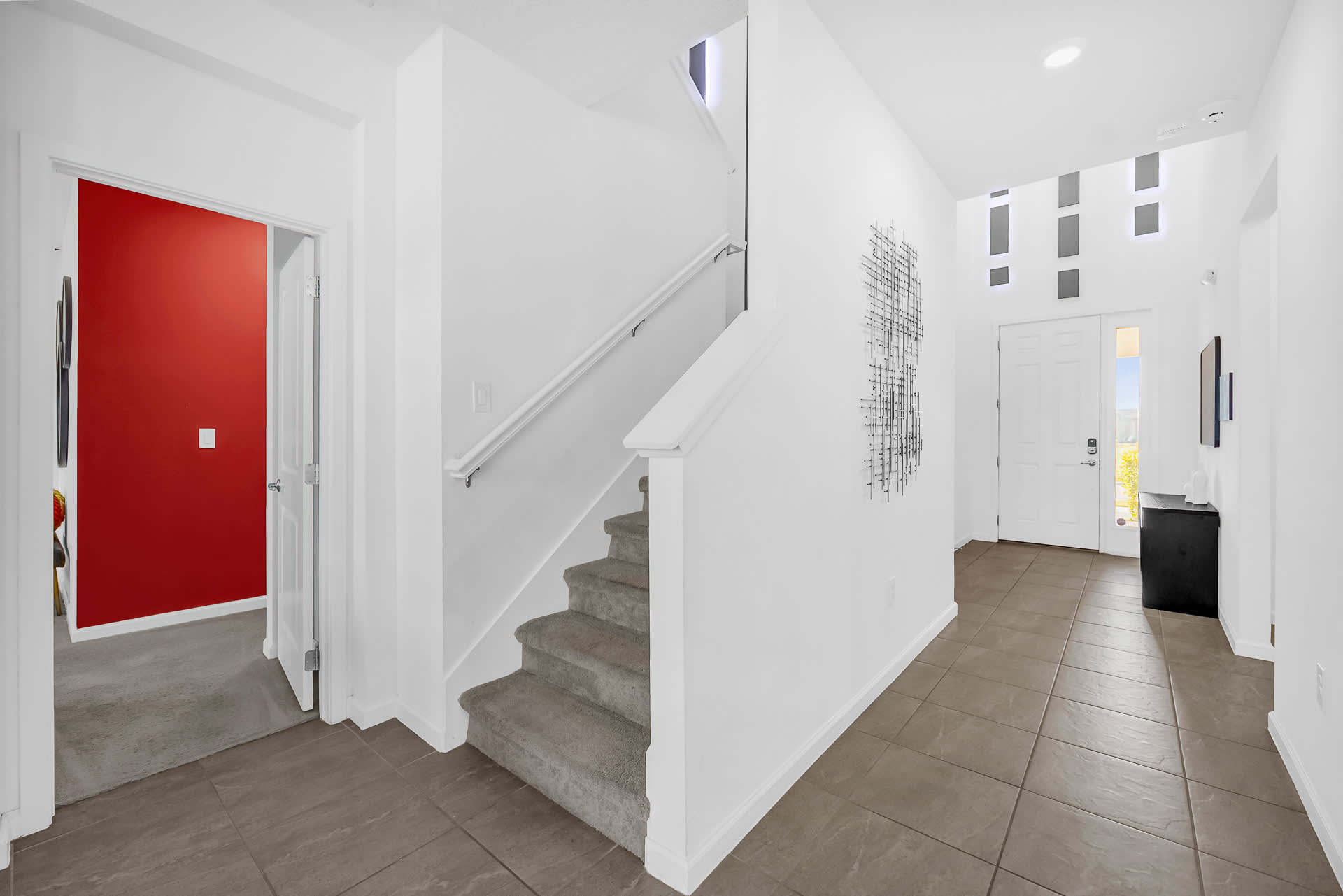 Bright foyer with tiled flooring, white walls, and a carpeted staircase leading upstairs. Modern wall art adorns one wall.