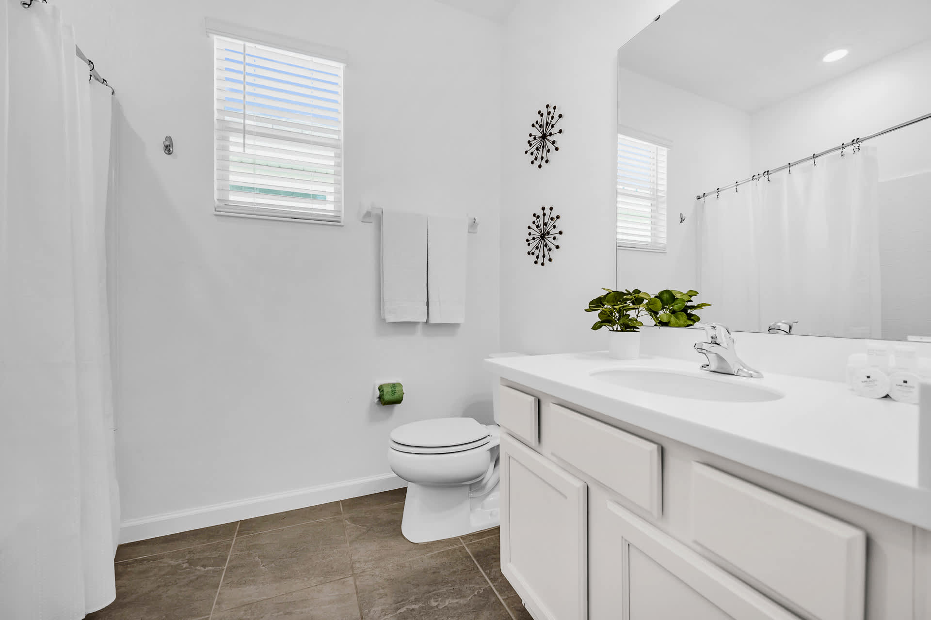 Bright white bathroom with a single vanity, toilet, and a shower-tub combo visible in the background. Two small windows provide natural light.