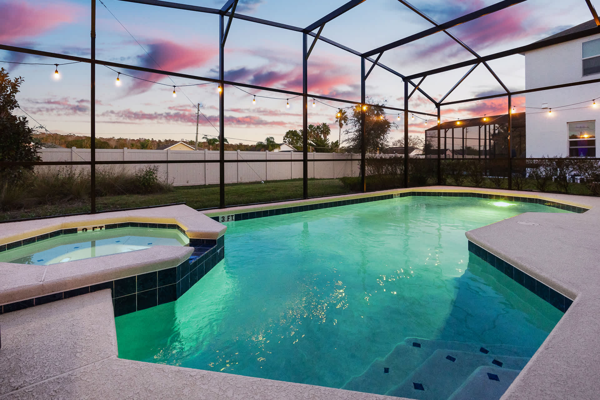 An outdoor screened-in pool area at dusk, showcasing a large swimming pool with an integrated hot tub, surrounded by concrete decking and string lights, with a distant view of the neighborhood and a vibrant sky.