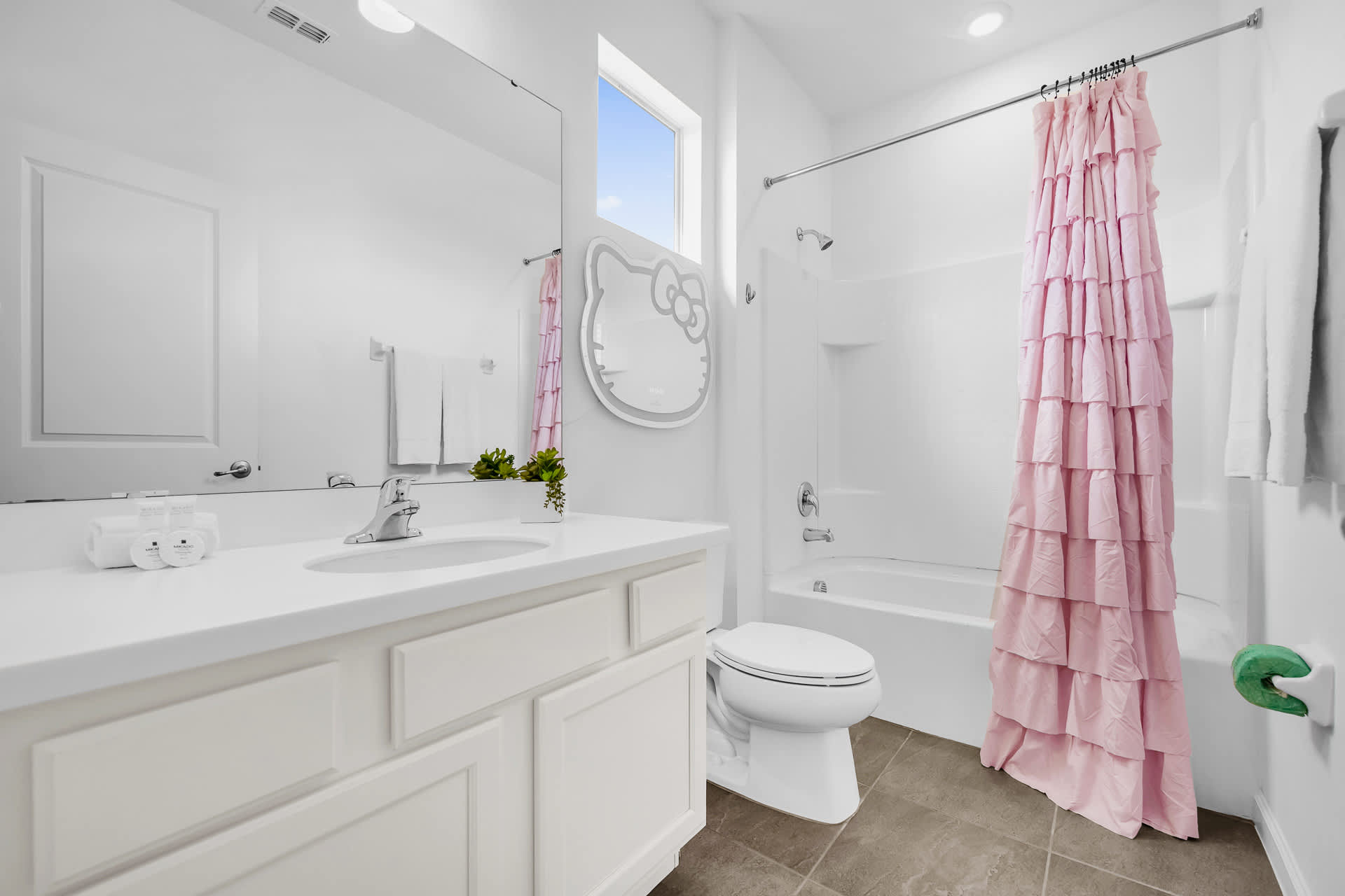Bright white bathroom with a single vanity, toilet, and a shower-tub combo featuring a ruffled pink shower curtain. A Hello Kitty shaped mirror with backlighting adds a playful touch.