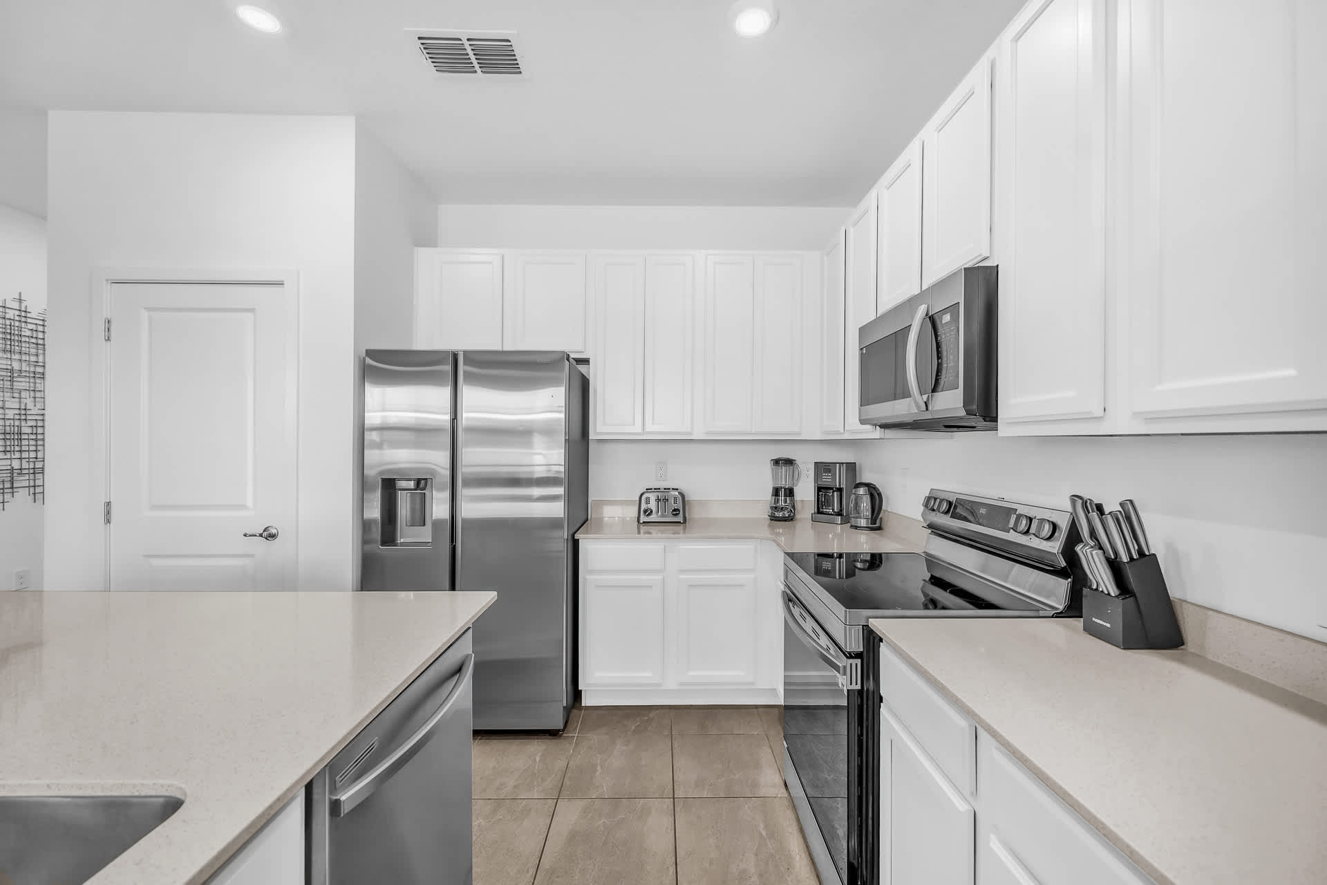 A well-equipped kitchen features white shaker cabinets, light countertops, and stainless steel appliances including a refrigerator, microwave, and electric range. A toaster and coffee maker are visible on the counter.