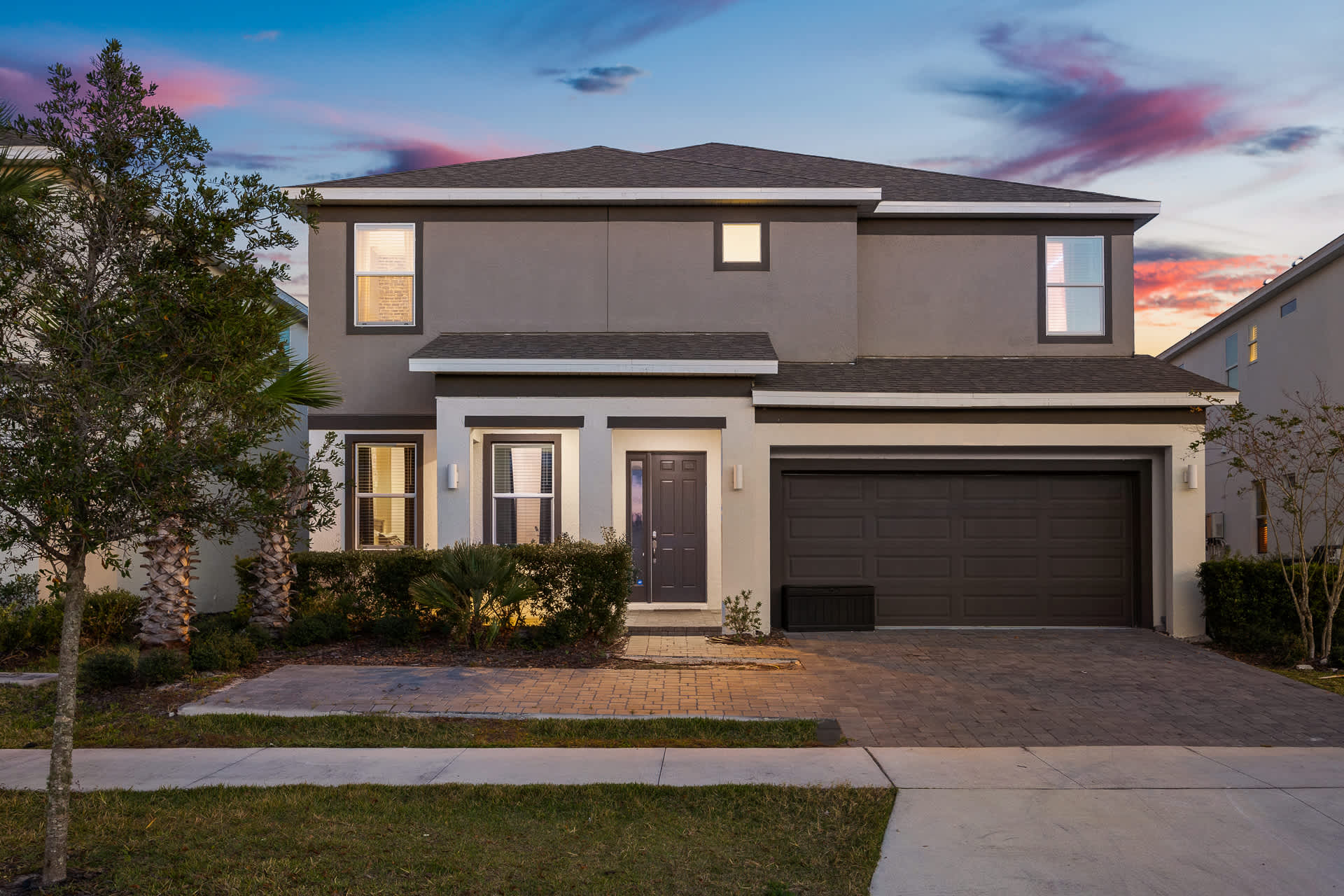 The front exterior of a modern two-story house with a gray and white facade, a two-car garage, and a paved driveway, surrounded by manicured landscaping and illuminated by exterior lights at dusk.