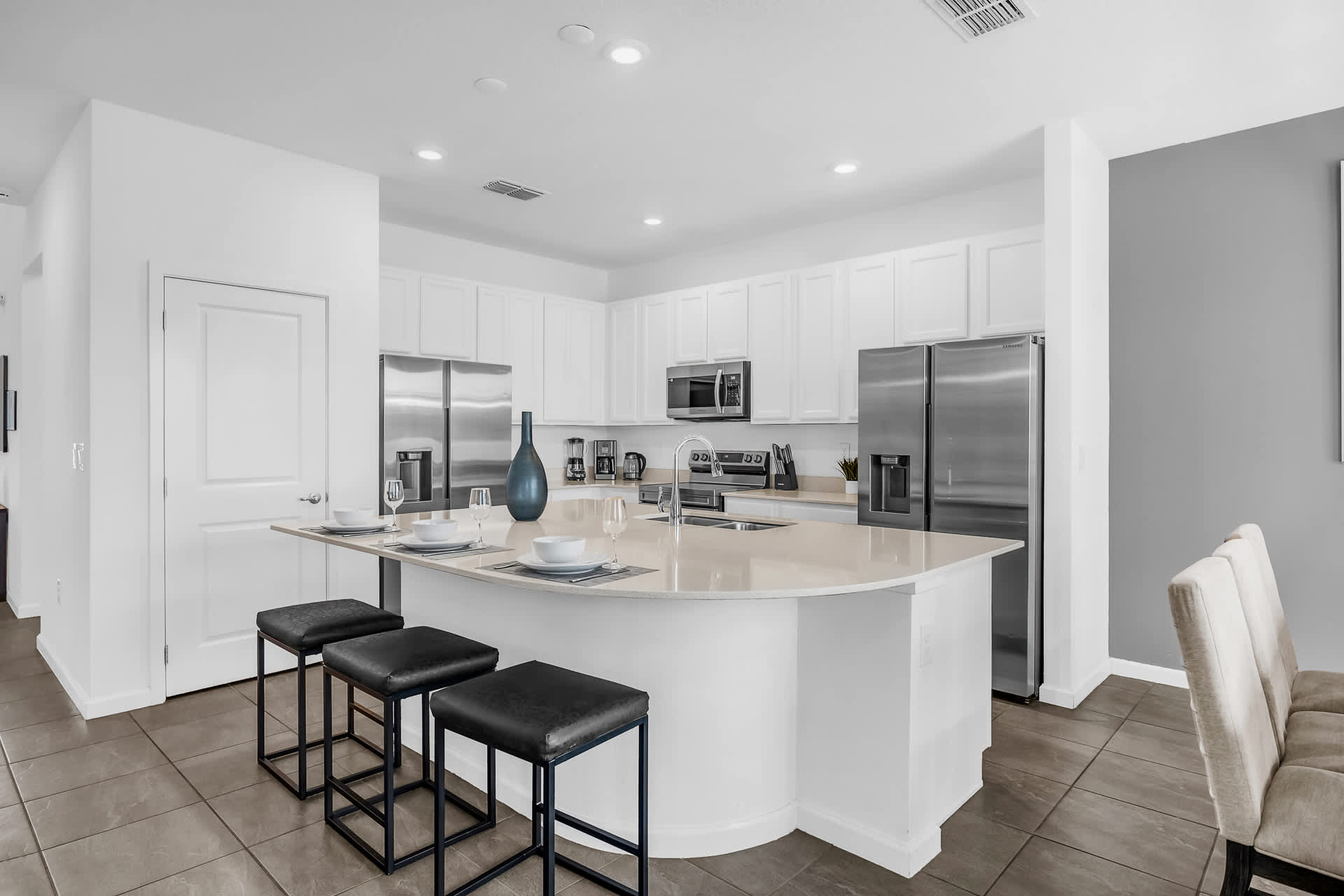 A modern kitchen features white shaker cabinets, stainless steel appliances including two refrigerators, and a large white quartz island with a sink and seating for three. The space is well-lit with recessed lighting.