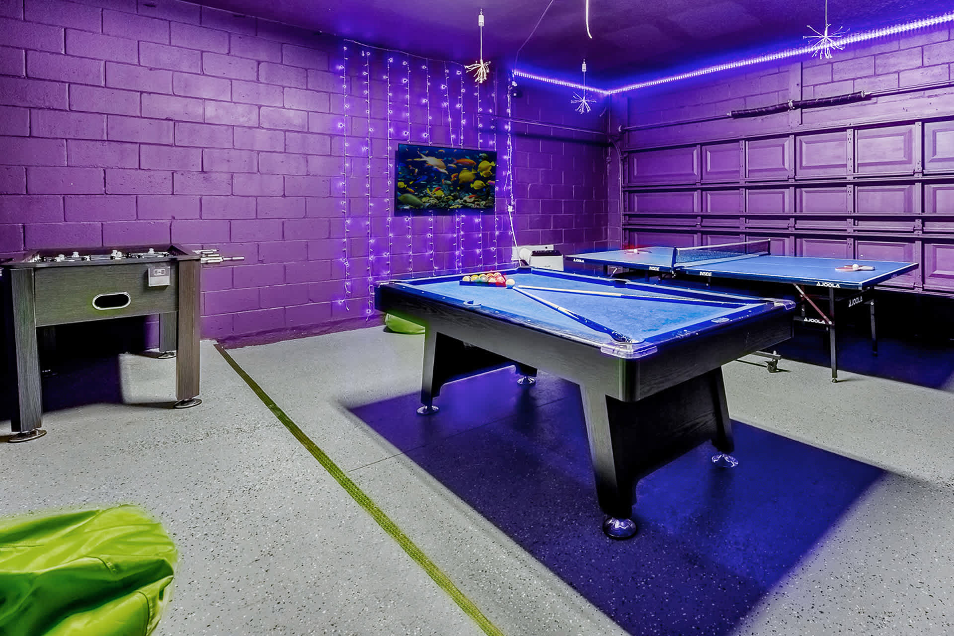 A game room with purple brick walls and purple LED lighting, featuring a pool table, a ping pong table, and a foosball table, with a wall-mounted TV displaying an aquarium scene.