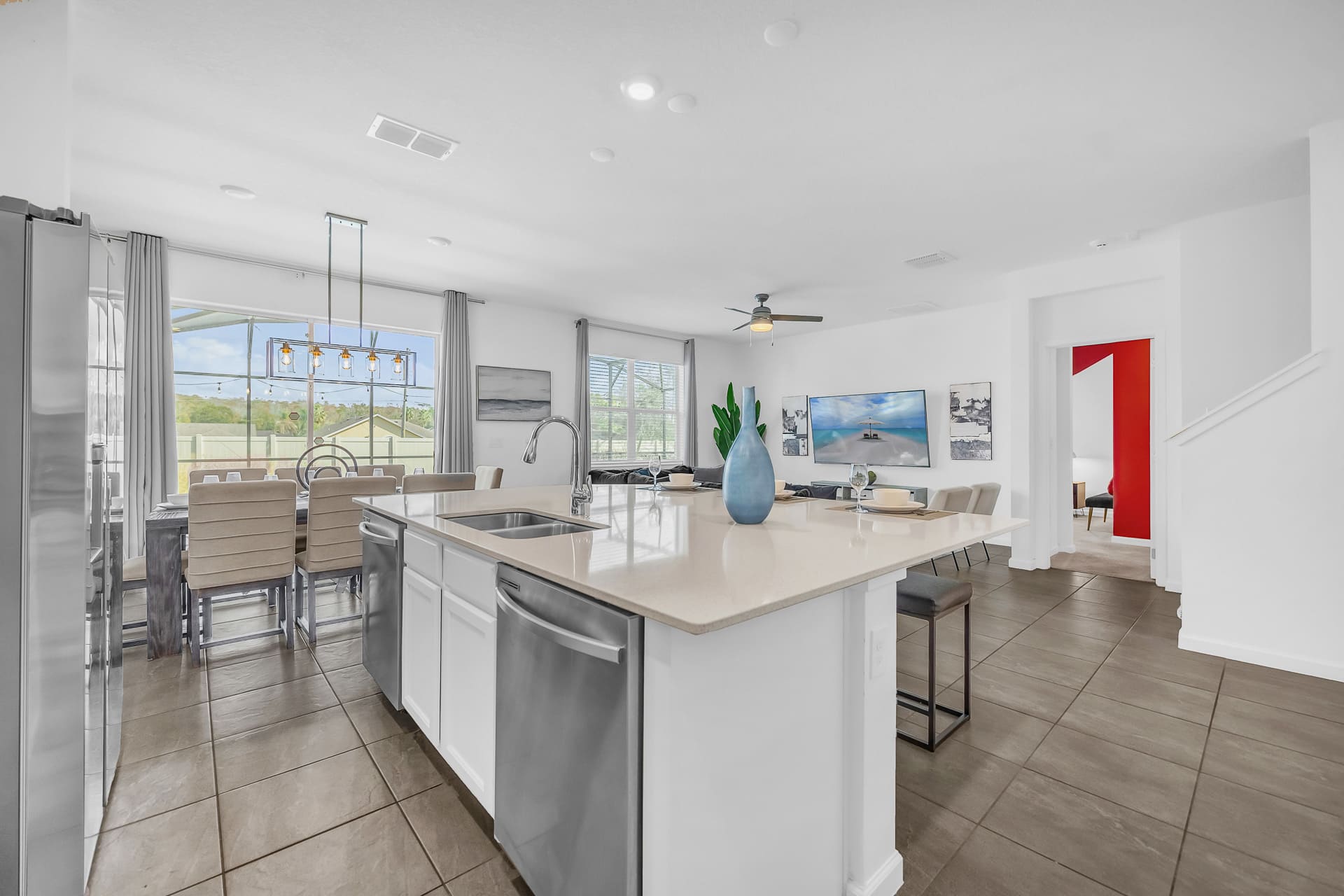 A bright, open-concept kitchen features a large white quartz island with a sink and bar seating, overlooking the dining area and outdoor pool through sliding glass doors. The kitchen is equipped with modern stainless steel appliances and white cabinetry.