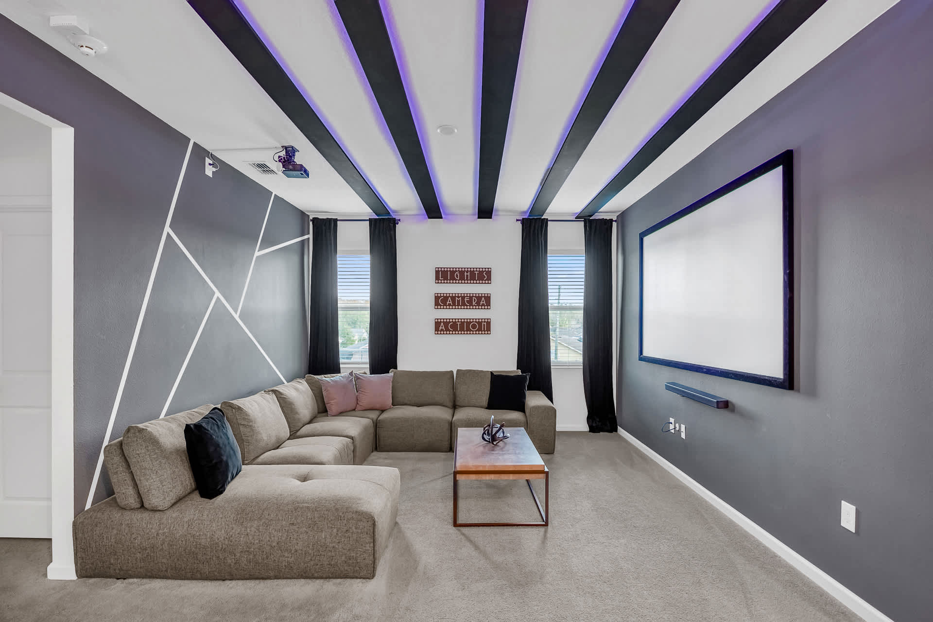 A modern media room with a large sectional sofa, a projector screen, and a projector, featuring a striking black and white geometric wall design and a ceiling with black beams and purple LED lighting.