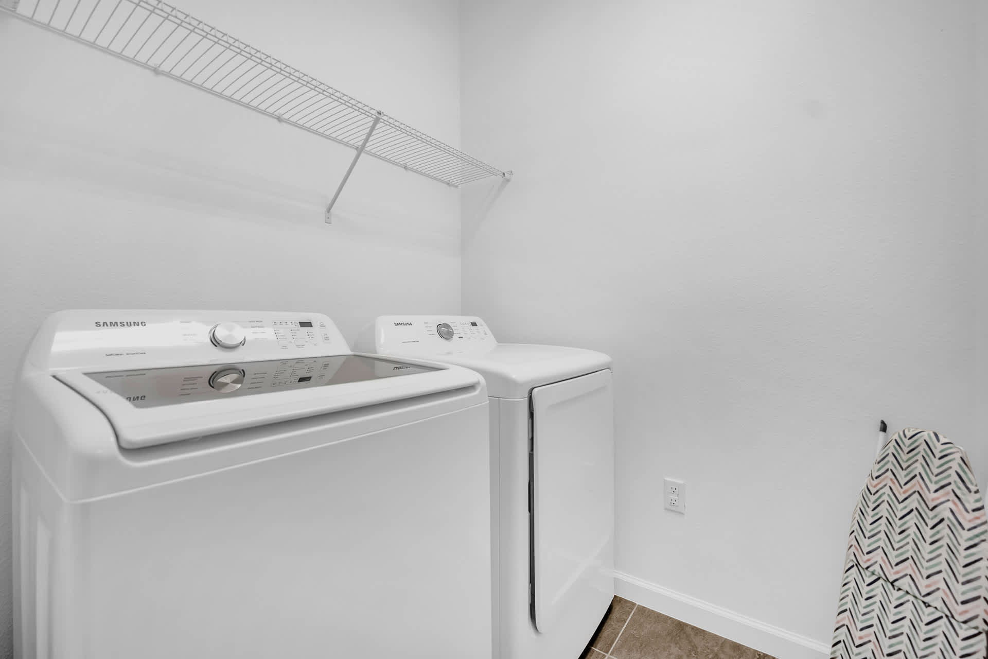 Functional laundry room with a white Samsung top-load washing machine and front-load dryer. A wire shelf is mounted above for storage, and an ironing board is visible.