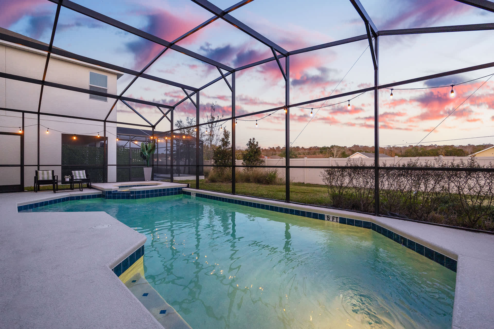 An outdoor screened-in pool area at dusk, featuring a large swimming pool with an integrated hot tub, surrounded by concrete decking and string lights overhead, with a vibrant sunset sky visible through the screen.