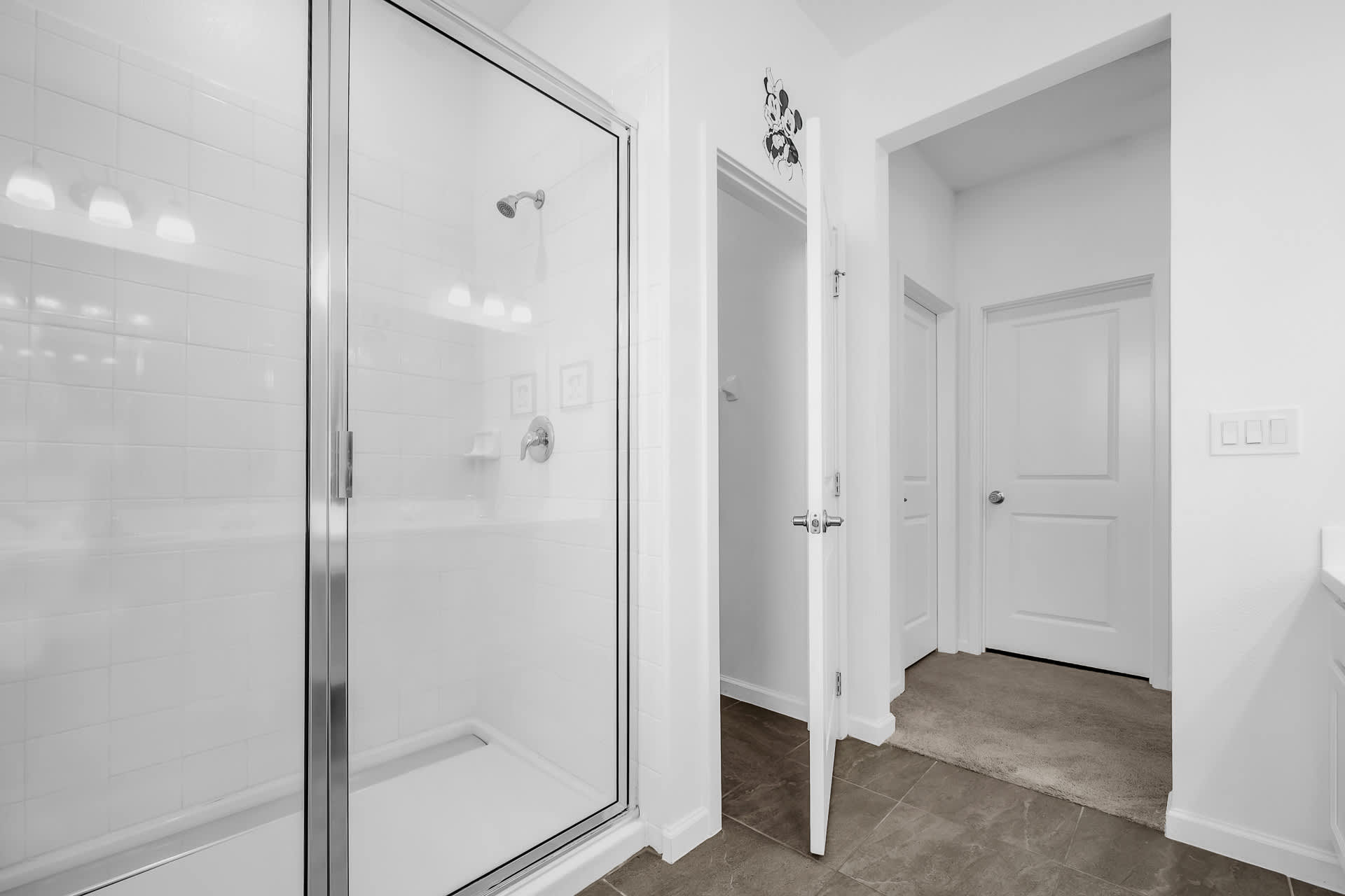 View of the bright bathroom's walk-in shower with white subway tiles and glass door, adjacent to a doorway leading to another room.