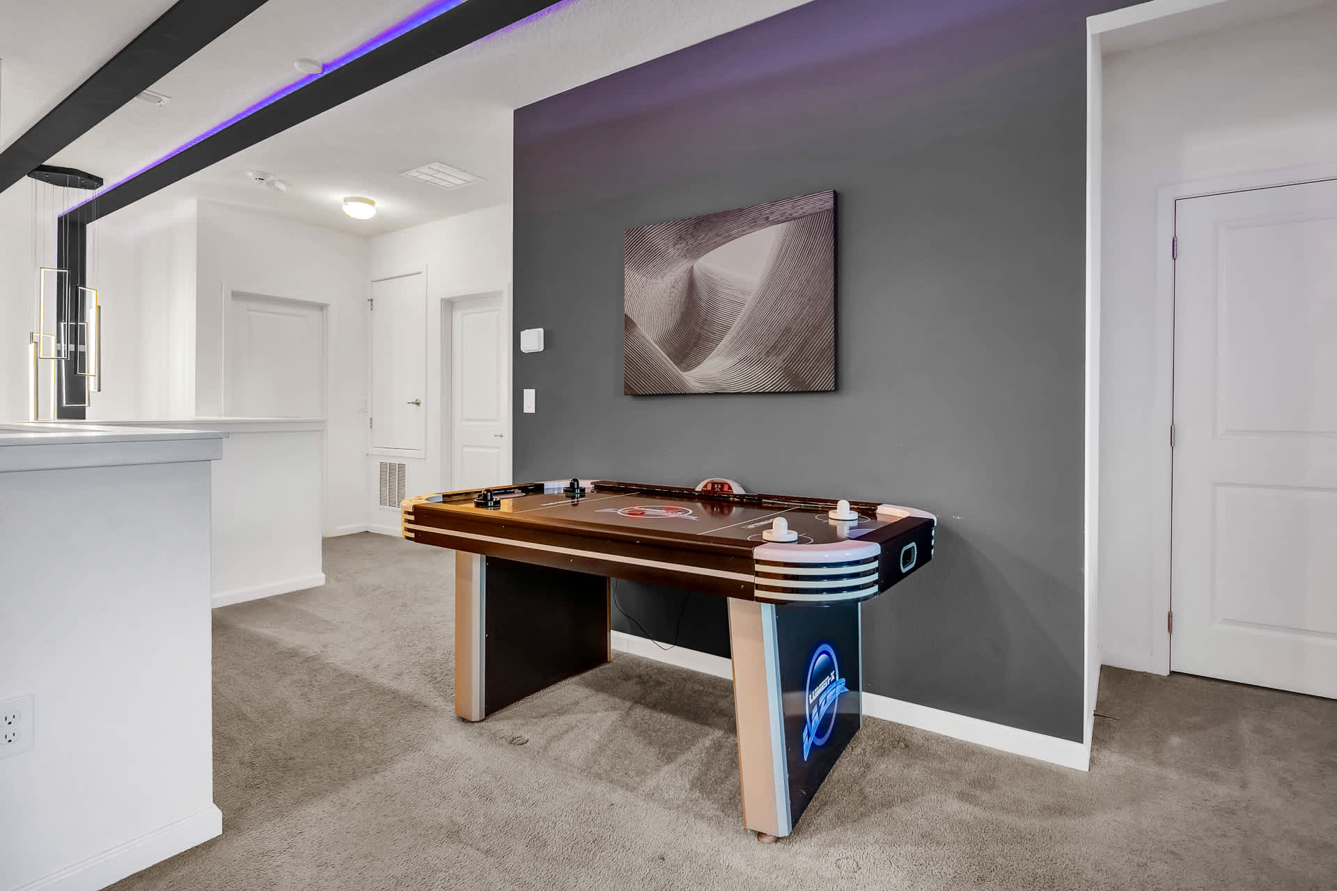 A vibrant air hockey table with illuminated sides is positioned against a dark gray accent wall featuring a textured abstract artwork. The room has light gray carpet and a ceiling with black beams and purple LED lighting.
