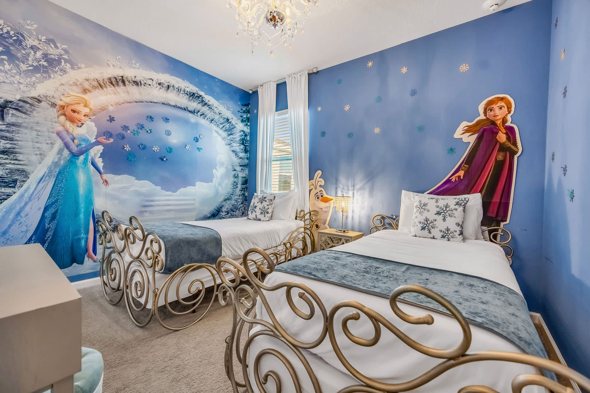 Frozen-themed bedroom with two ornate gold twin beds and a large mural of Elsa and an icy landscape. The blue walls are adorned with snowflake decals, and a small Olaf figure stands between the beds.
