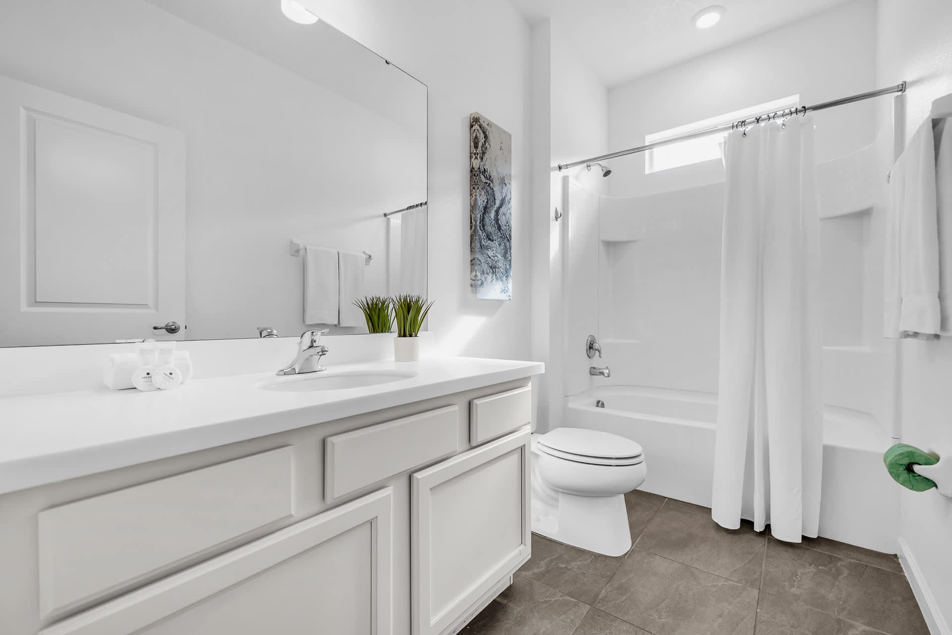 Clean, modern white bathroom with a single vanity, toilet, and a shower-tub combo with a plain white curtain. A large mirror and simple green plant decor complete the minimalist look.