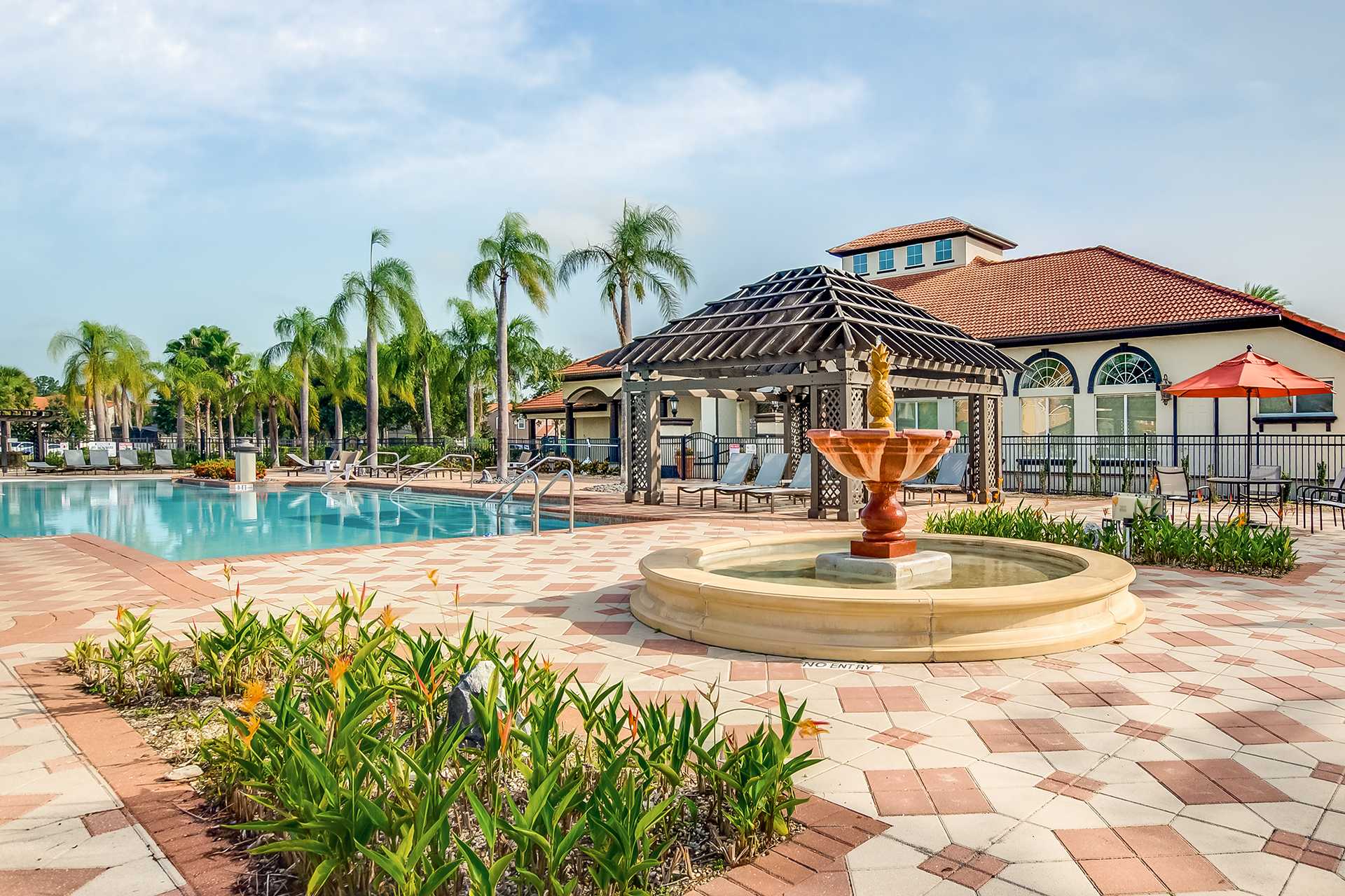 A wide view of the communal pool area featuring a multi-tiered fountain with a golden statue, surrounded by a patterned brick patio and tropical plants. The large swimming pool, a wooden pergola, and the clubhouse are visible in the background.