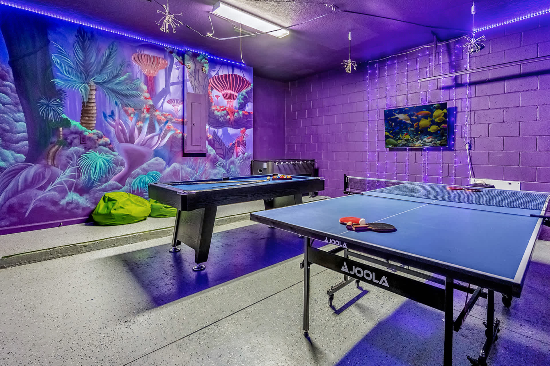 A vibrant game room with purple walls, featuring a pool table, a foosball table, and a ping pong table, illuminated by purple LED strip lighting and a colorful jungle-themed mural on one wall.