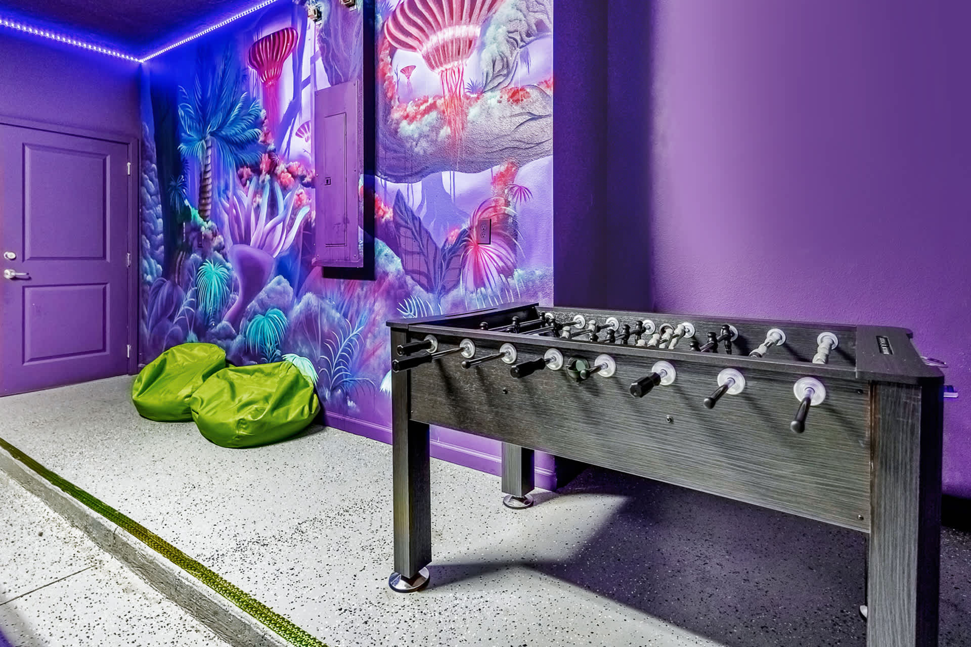 A vibrant game room with purple walls and a colorful jungle-themed mural, featuring a foosball table and two green bean bag chairs, with purple LED strip lighting illuminating the space.
