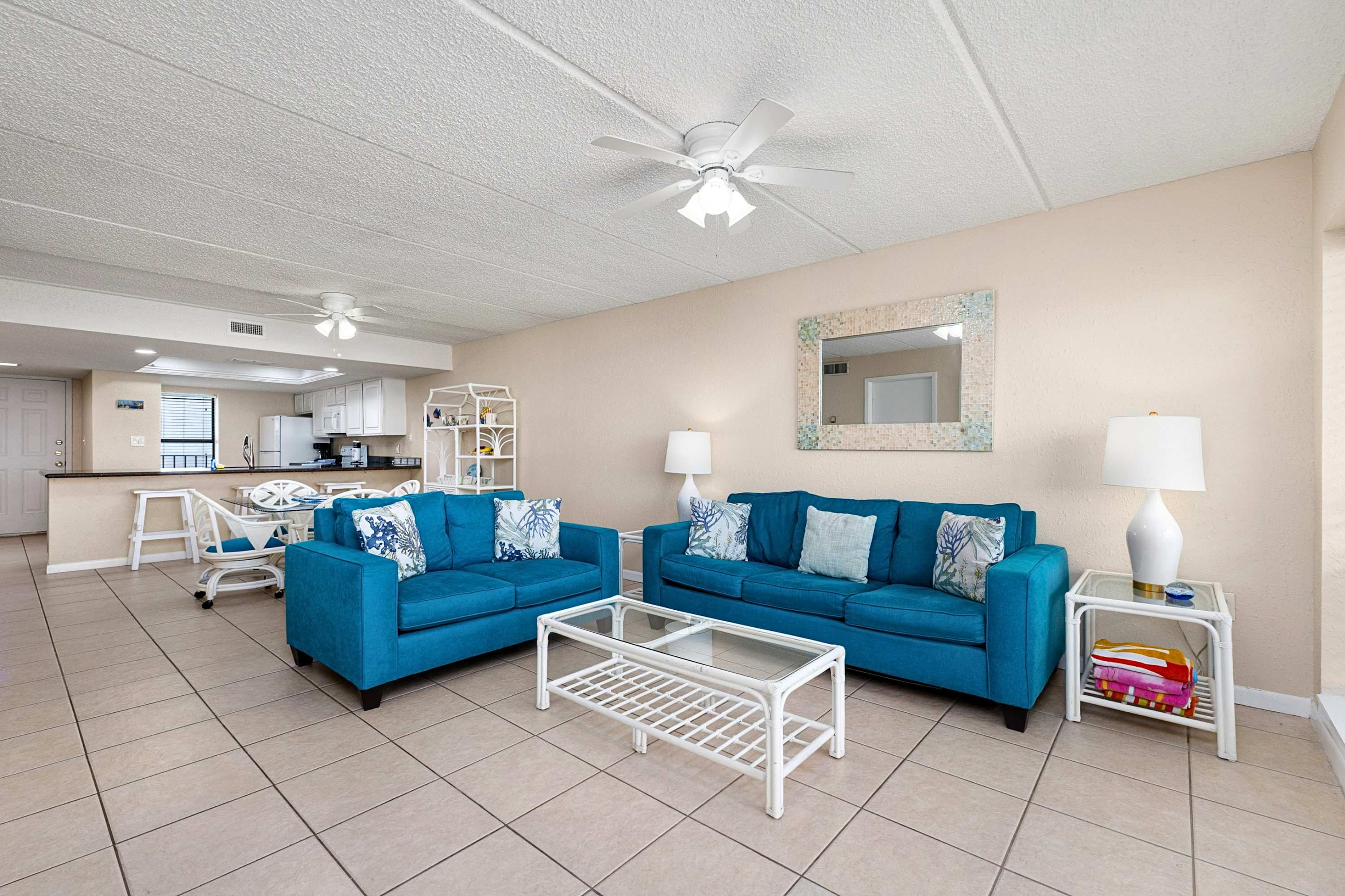 Open-concept living space with two blue sofas, a glass coffee table, and a dining area with a glass-top table and white wicker chairs, leading into a kitchen with a breakfast bar.