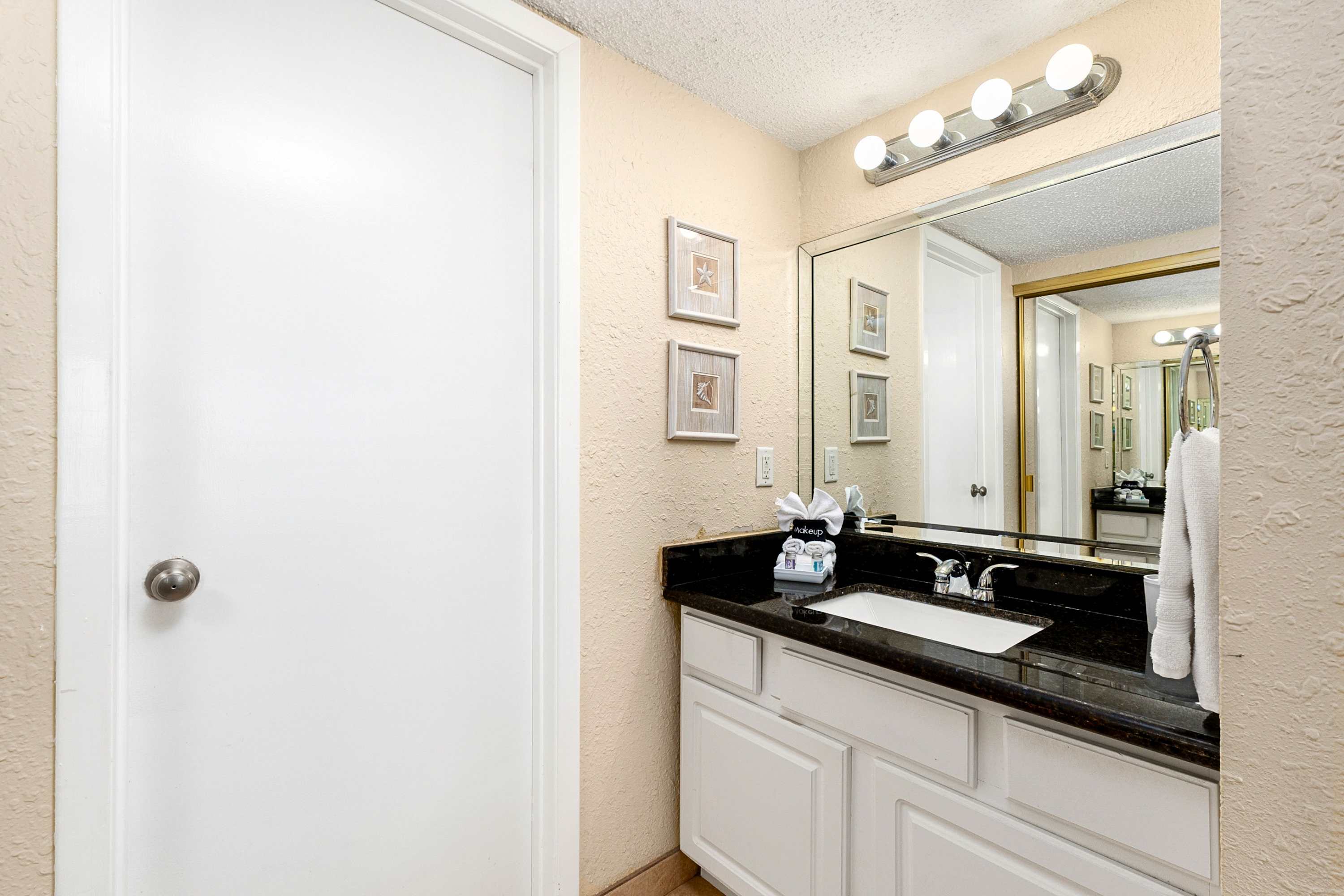 A bathroom vanity area featuring a dark granite countertop with a white sink, a large mirror, a three-bulb vanity light, and two framed seashell artworks on the light-colored wall.