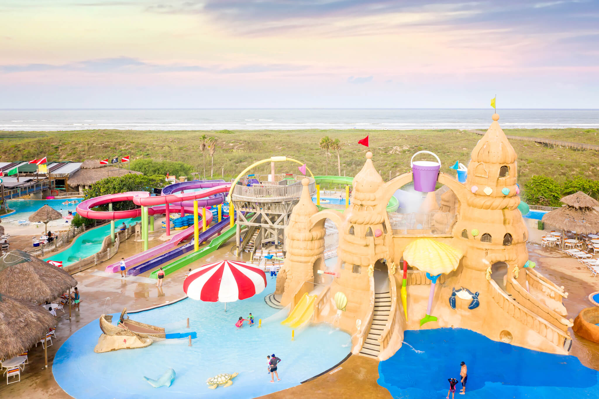 Aerial view of a vibrant water park featuring a large sandcastle play structure with slides and a dumping bucket, multiple colorful water slides, and various pools. The ocean and a sandy beach are visible in the background under a soft, cloudy sky.